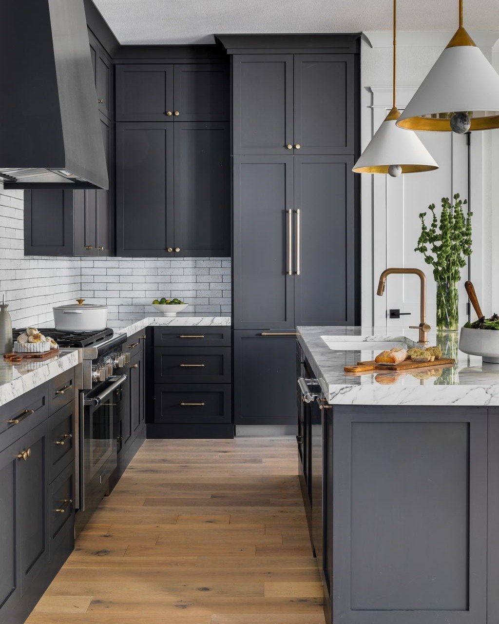 A modern kitchen with deep charcoal gray cabinets, white marble countertops, white subway tile, and light wood floors.