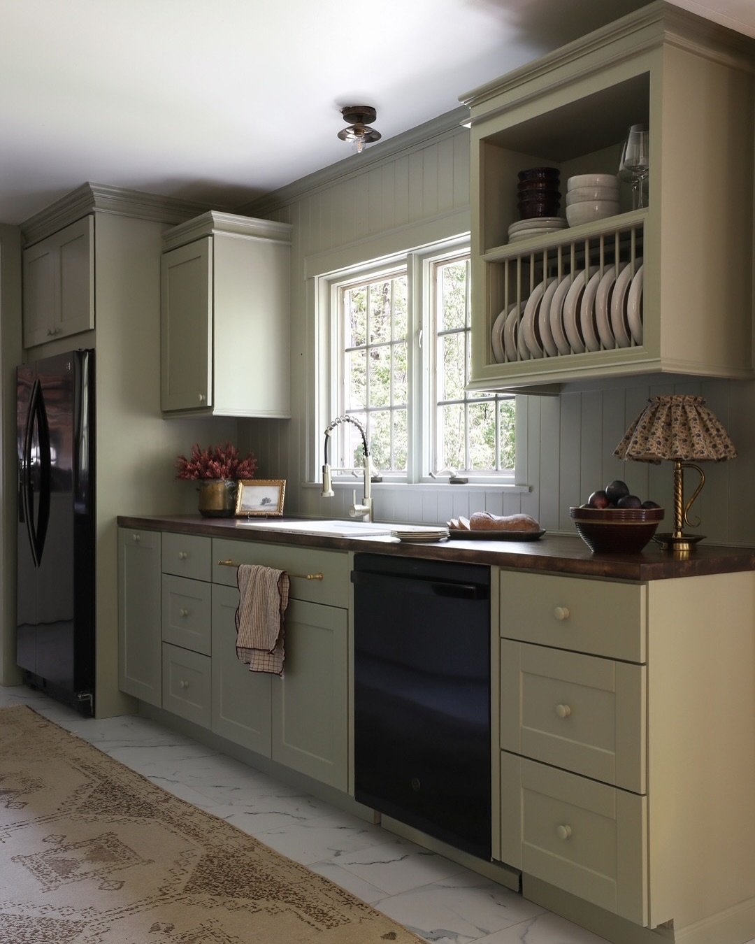 A cozy kitchen with muted sage green shaker-style cabinets, dark wood countertops, and a white marble floor.