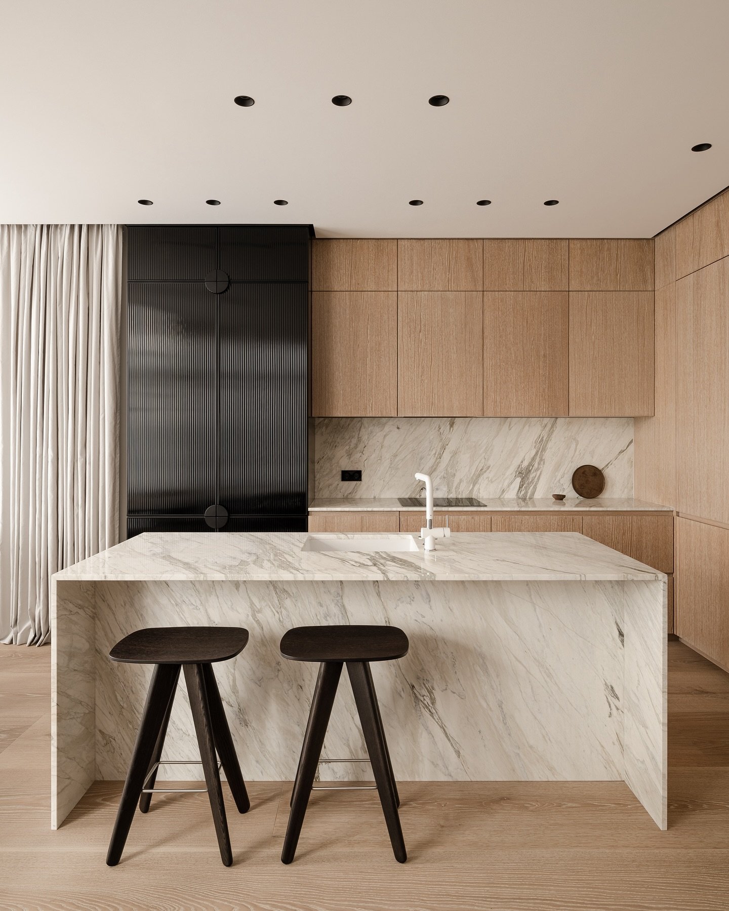 A minimalist kitchen with light wood grain cabinets, a waterfall marble island, and a fluted black cabinet unit.