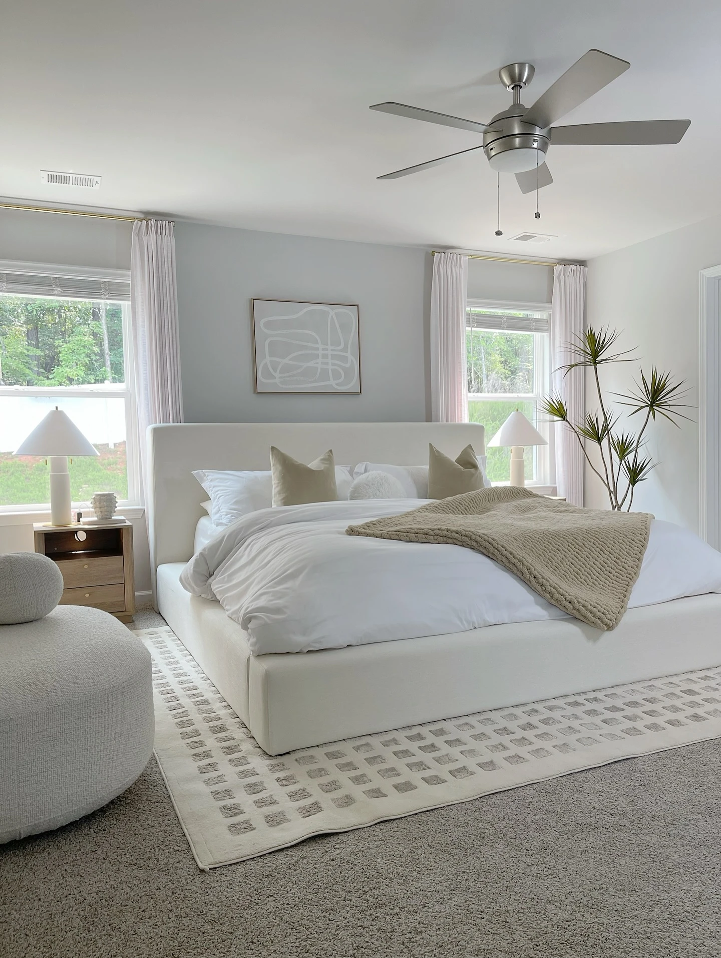 Cozy small bedroom with white bed, ceiling fan, sheer curtains, nightstand, and potted plant.