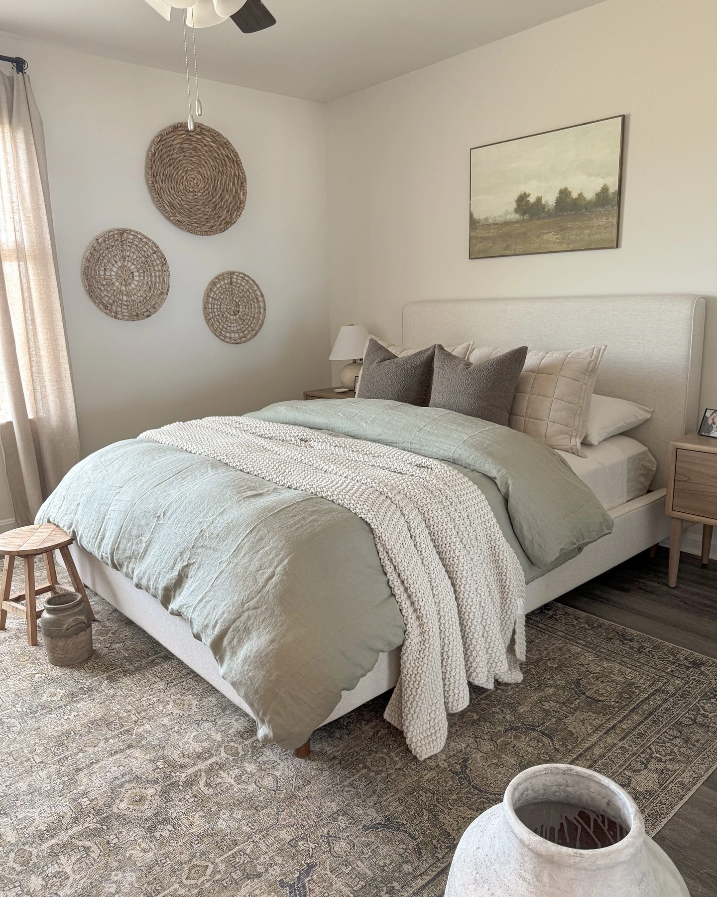 Rustic small bedroom with beige bed, sage green duvet, woven baskets, ceiling fan, and ceramic vase.