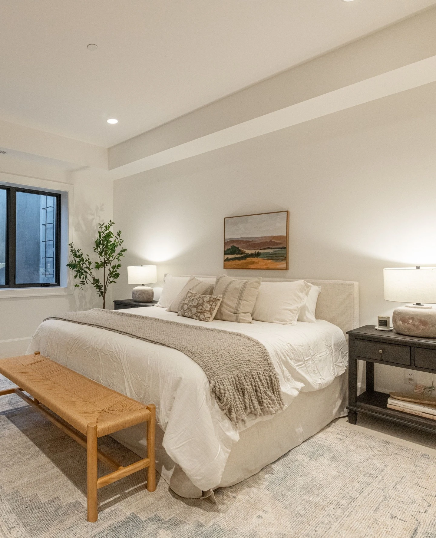 Serene small bedroom with white bed, wooden bench, landscape painting, potted plant, and area rug.
