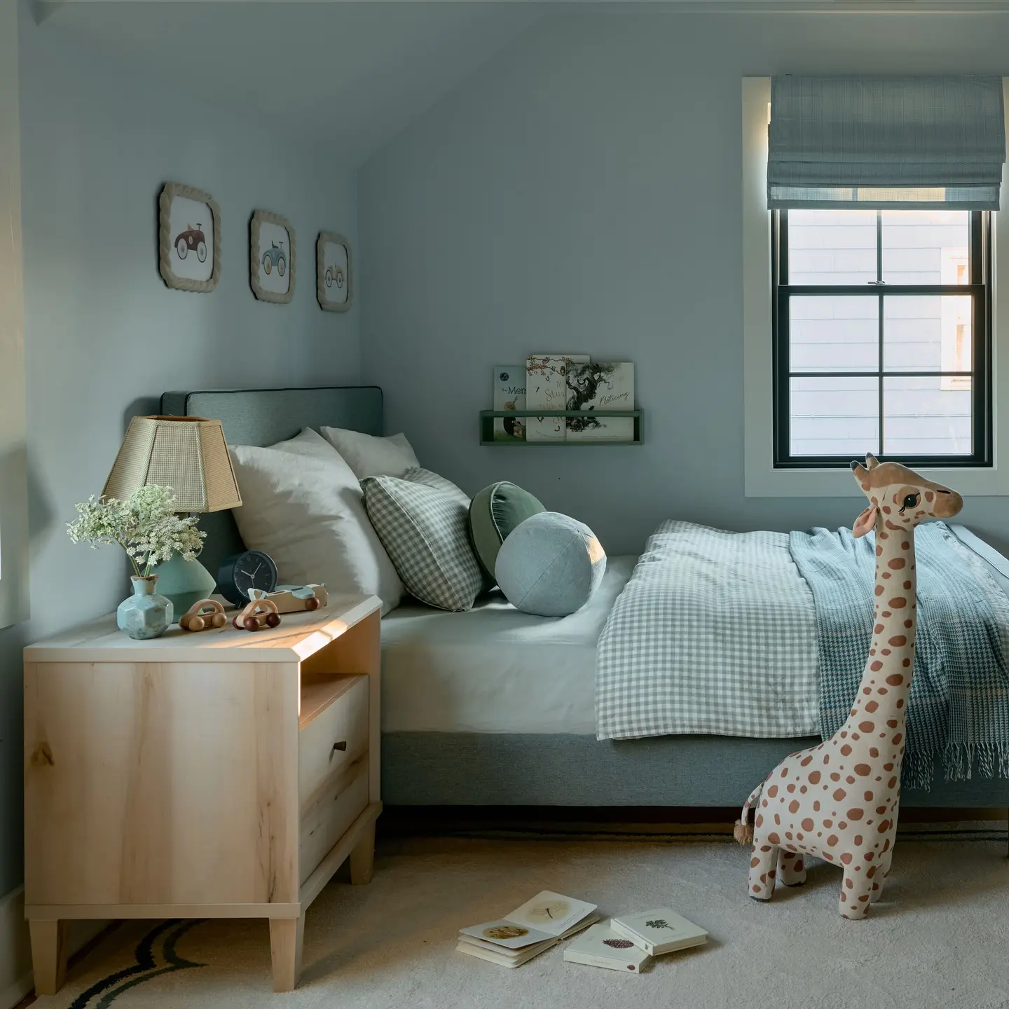A Kid's bedroom with a single bed dressed in light blue and white gingham bedding. The walls are a soft, dusty blue. A large giraffe stuffed animal stands at the foot of the bed, and a light wood nightstand with a lamp sits beside it. A small, black-framed window is covered with a blue Roman shade.