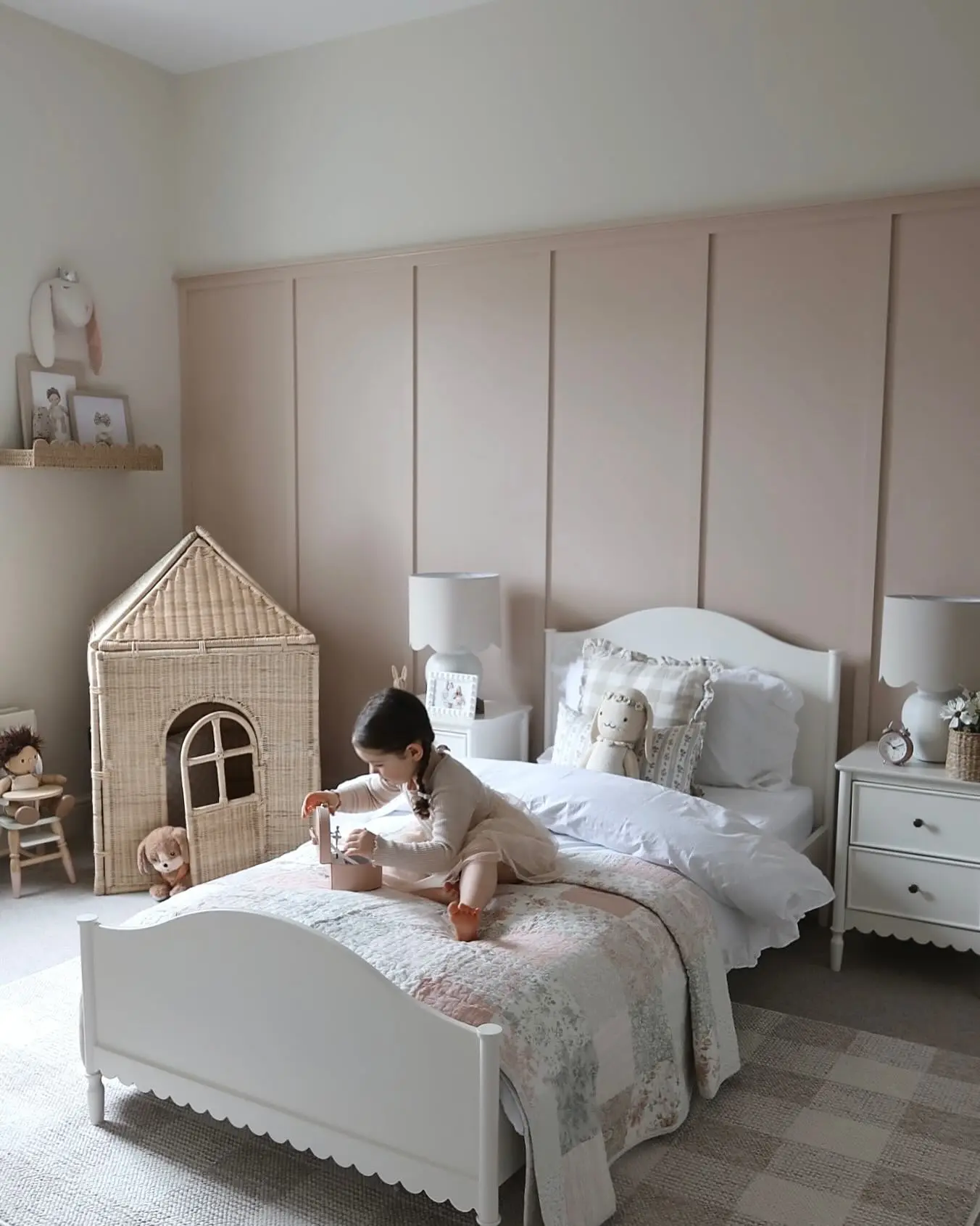 A little girl is playing on a white, scallop-edged bed in a bedroom with a blush pink board-and-batten accent wall. The room is decorated with a wicker playhouse, a checked rug, and soft, neutral furnishings. Two white nightstands with matching lamps flank the bed.