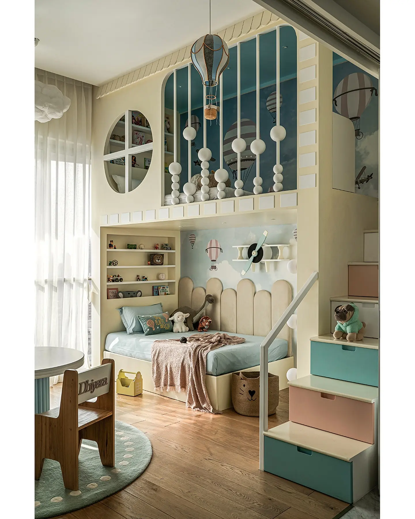 A child's bedroom featuring a two-story loft structure with an adventure and hot air balloon theme. The lower level has a single bed with a light wood headboard and built-in shelves. The upper loft is decorated with a mural and a hot air balloon lamp hangs from the ceiling. Steps with colorful drawers lead to the top level.