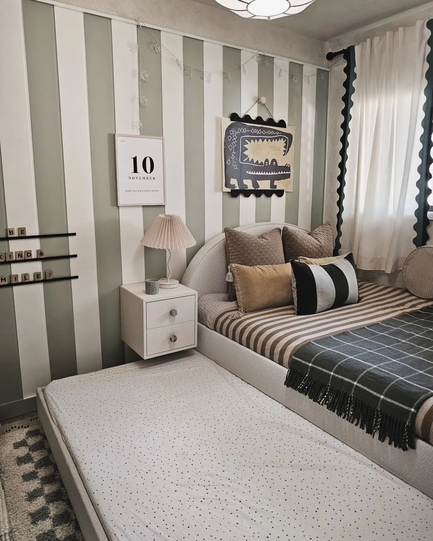 A Kid's bedroom with a single bed that has a pull-out trundle bed underneath, perfect for sleepovers. The room features a wall with alternating stripes of light gray and white, a small white nightstand with a pleated lamp, and an eclectic mix of bedding and wall art.