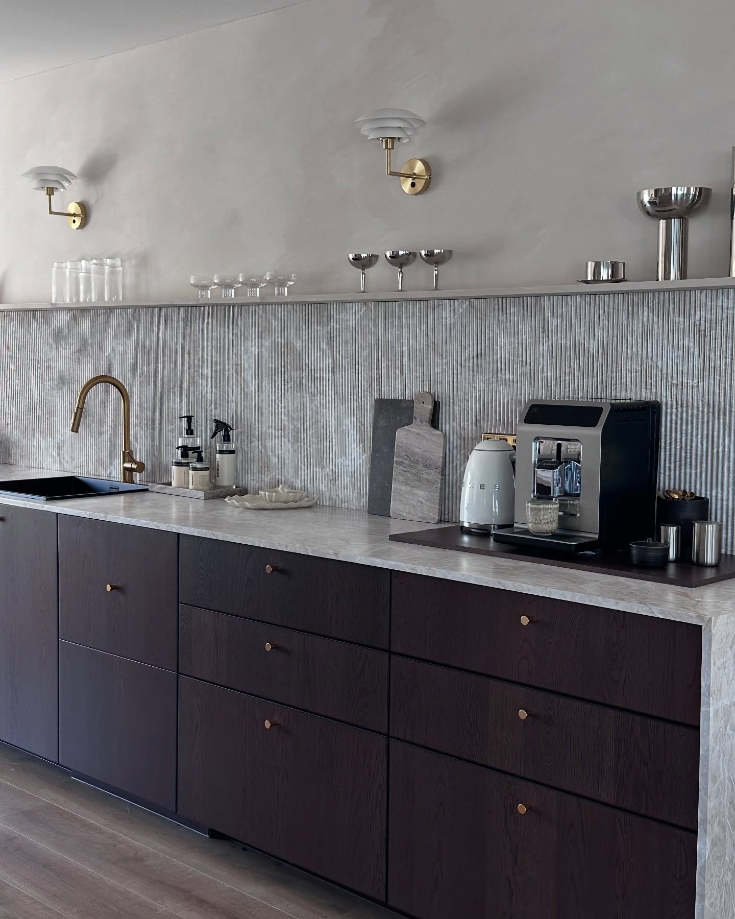 A contemporary kitchen featuring a grooved, textured backsplash in a muted gray tone, contrasting with dark wood cabinets and a white marble countertop.