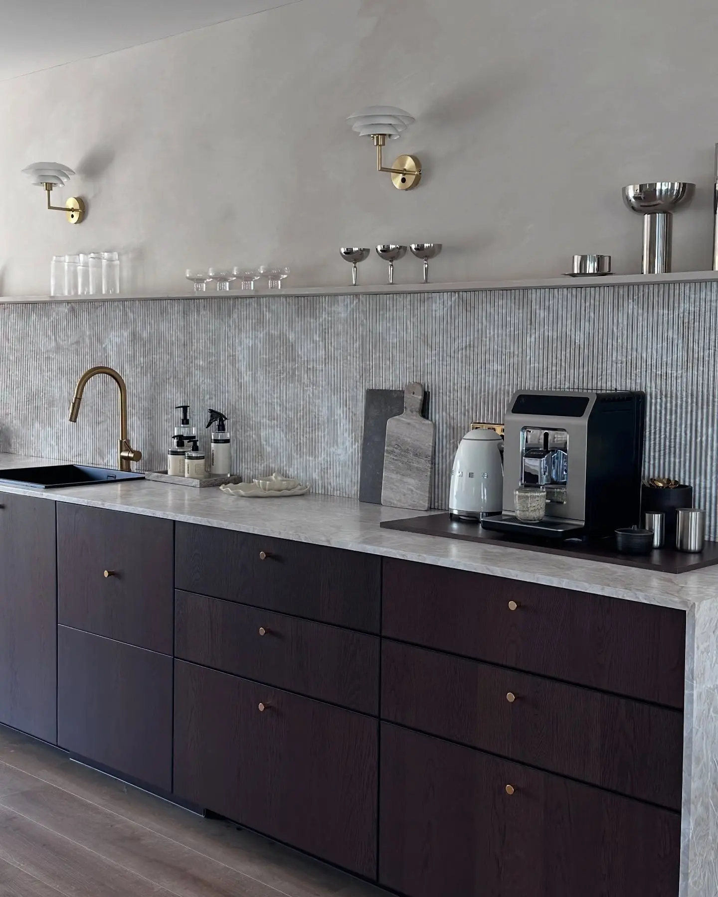 A contemporary kitchen featuring a grooved, textured backsplash in a muted gray tone, contrasting with dark wood cabinets and a white marble countertop.
