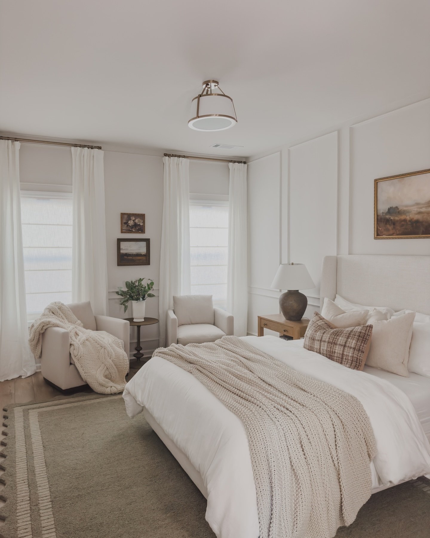 A cozy and elegant guest bedroom featuring a queen-sized bed with white linens and a chunky knit throw, two comfortable armchairs, and minimalist decor in a neutral color palette.