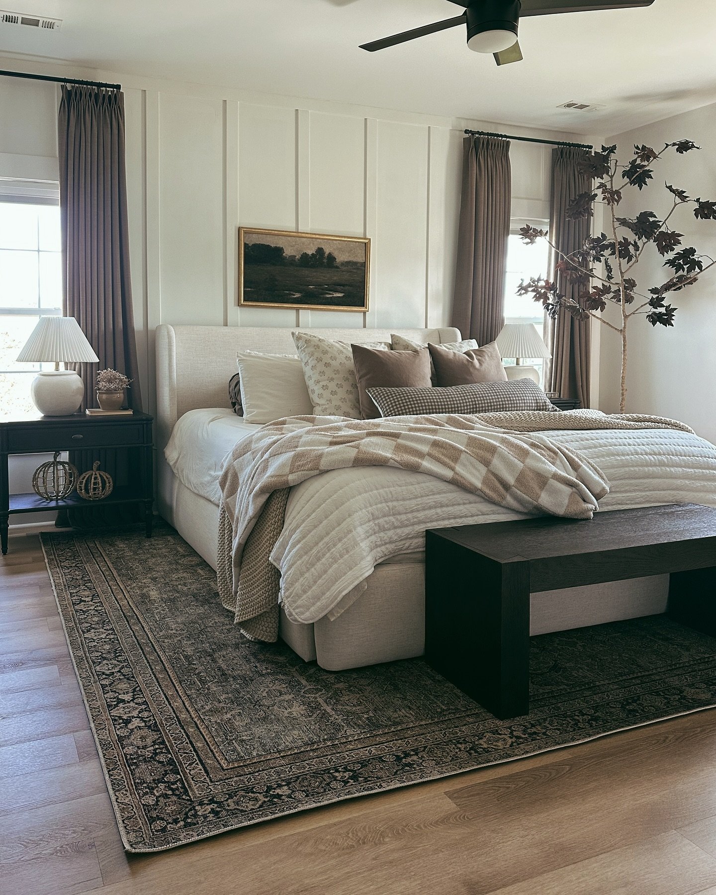 A neutral-toned bedroom with a paneled accent wall, dark wood nightstands, and a bed layered with textured pillows and blankets. A vintage-style rug covers the light wood floor, and a dark bench sits at the foot of the bed.