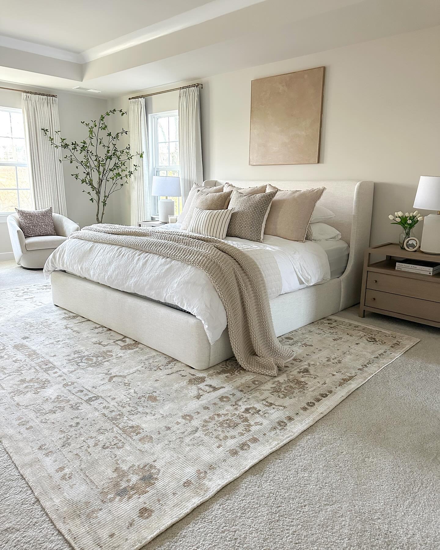 A neutral bedroom with a large, light-colored patterned rug placed under a white upholstered bed with beige pillows and throws.