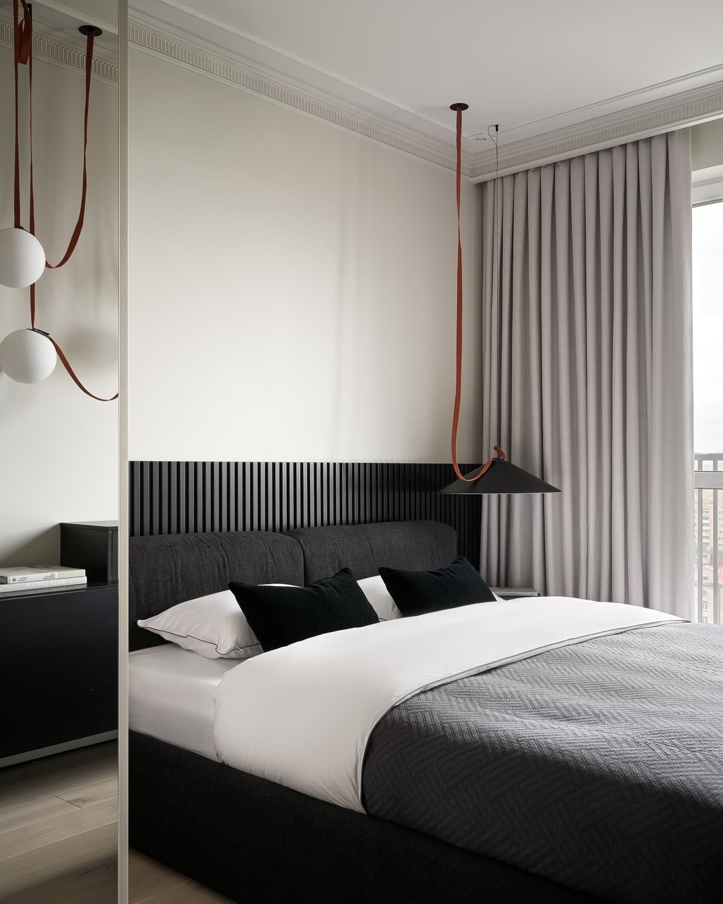 A contemporary black and white bedroom featuring a slatted black headboard, a plush bed with white and gray bedding, a modern hanging pendant lamp, and light gray curtains.