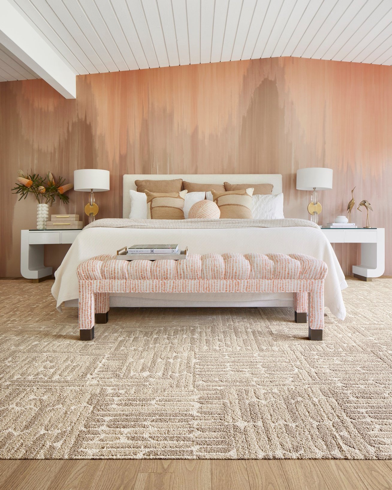 A large, light-colored abstract rug with a geometric pattern in beige and cream is placed under a bed and a bench with pink patterned upholstery.