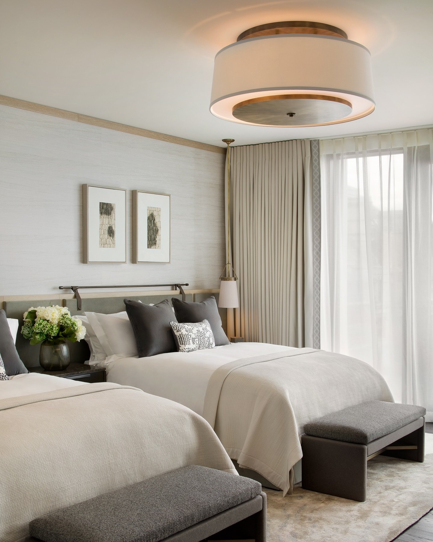 A modern twin guest bedroom with two beds featuring gray and white bedding, dark gray pillows, and upholstered gray benches at the foot of each bed. The room has textured white walls, a large drum pendant light, and framed abstract art.