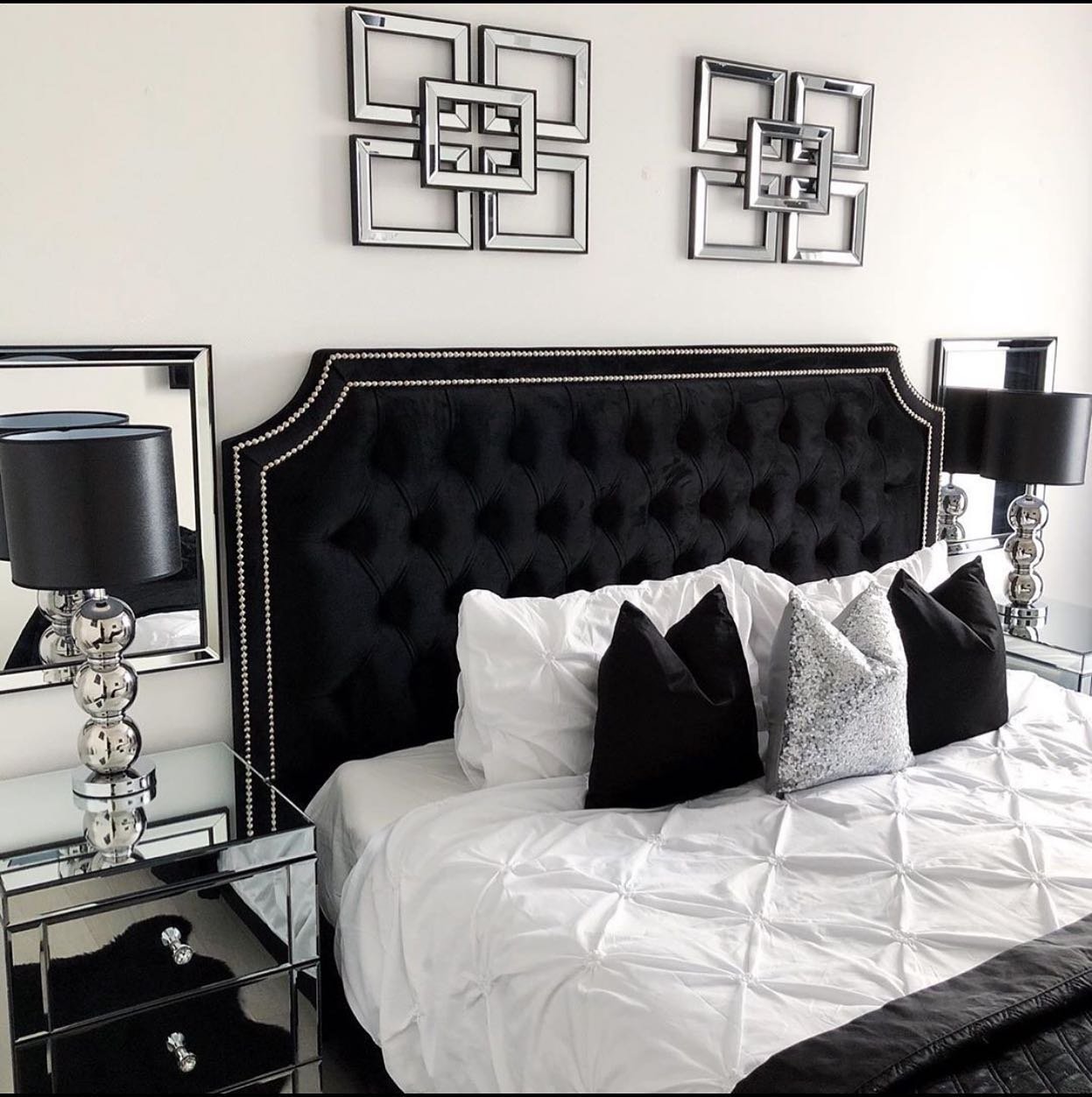 A glamorous bedroom with a black tufted velvet headboard, mirrored nightstands with black lamps, white bedding, and silver geometric wall art.
