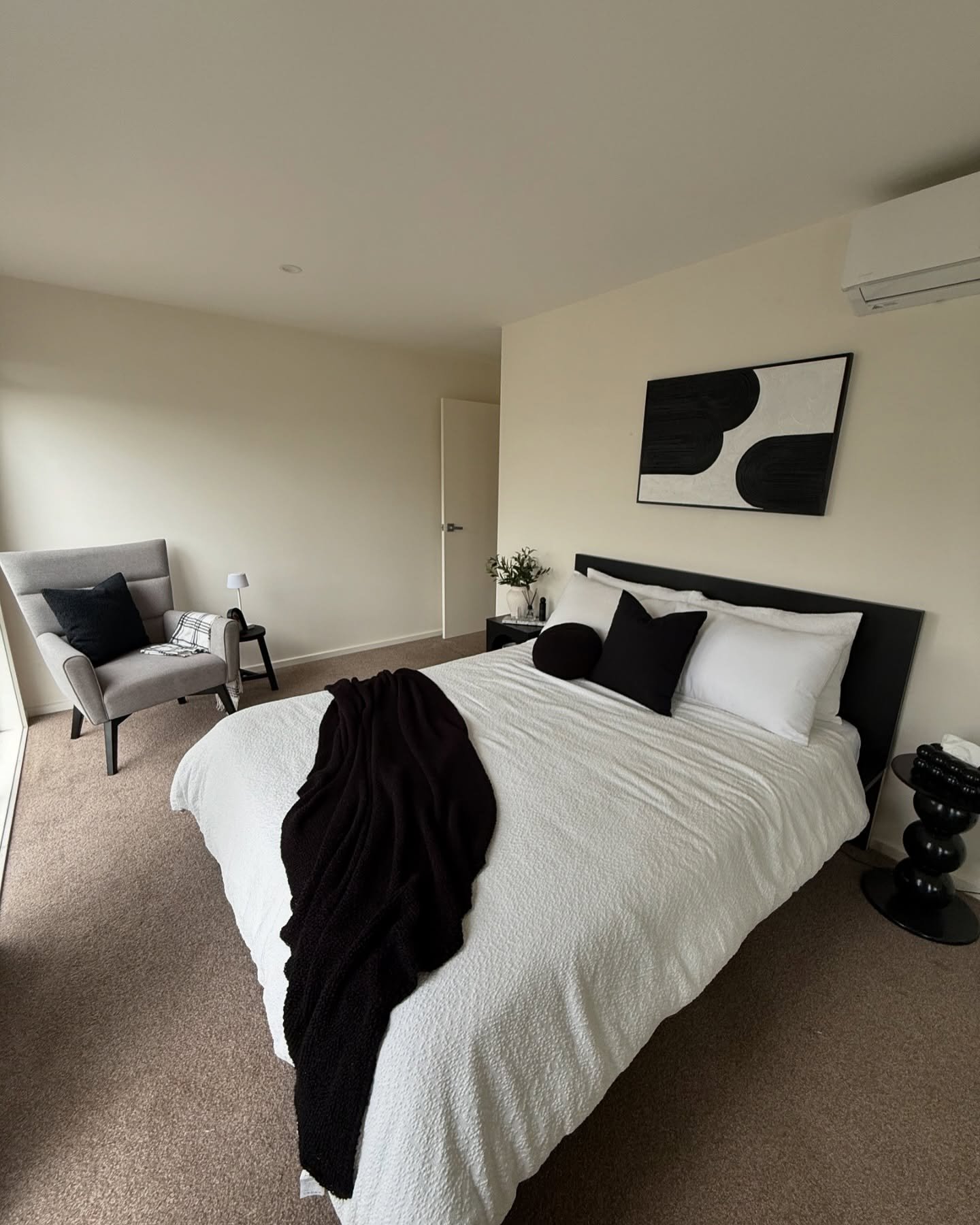 A modern and minimalist bedroom with a black and white color scheme, featuring a textured white bed, an abstract art piece, and a gray accent chair with a black cushion.