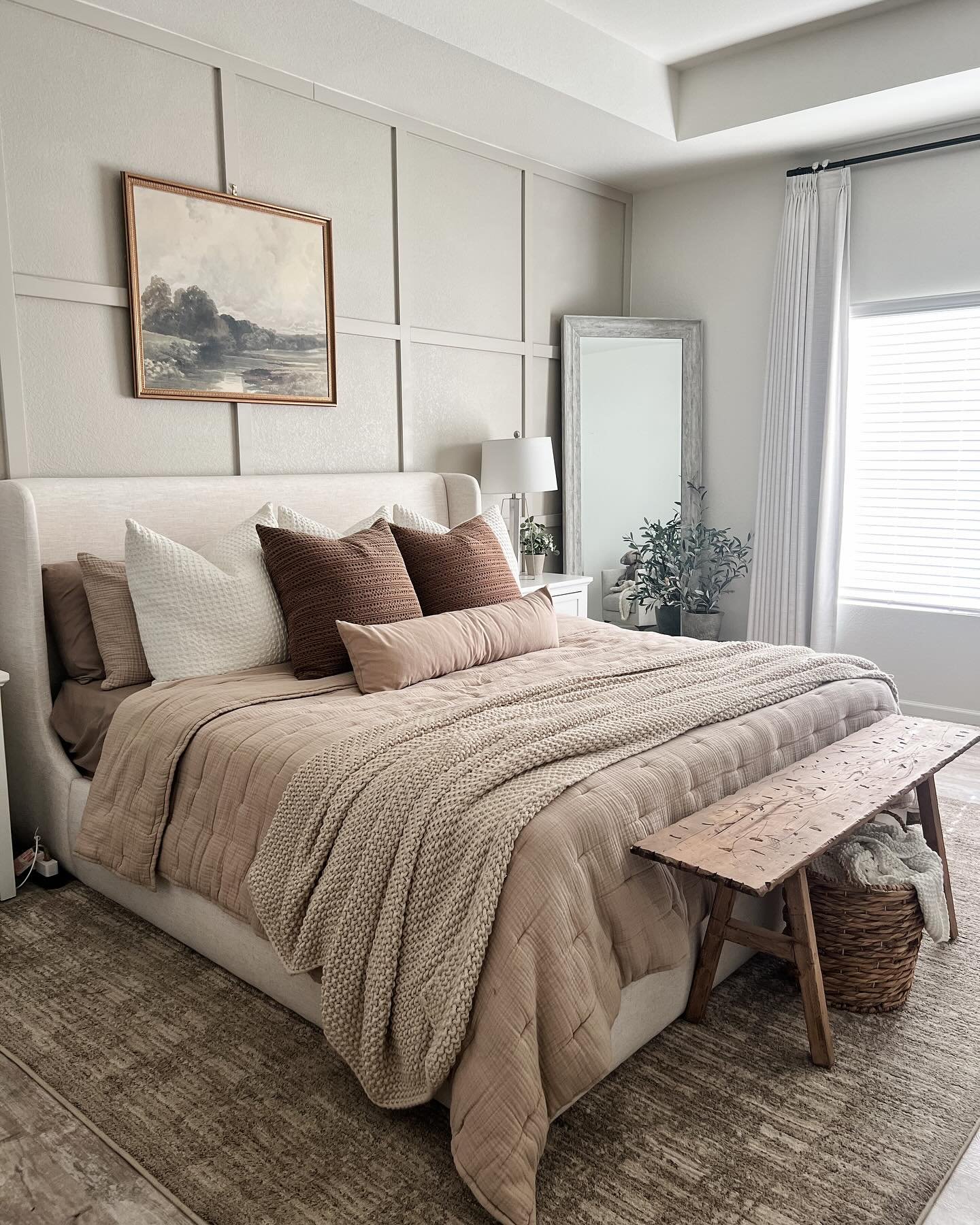 A cozy guest bedroom with an upholstered bed, a neutral color palette, and a paneled accent wall. The bed is layered with soft brown and cream bedding, a chunky knit blanket, and a rustic wooden bench at the foot.