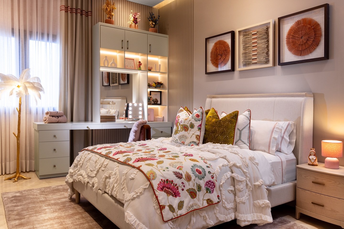 An eclectic bedroom with a floral comforter, a white tufted bed frame, a lit vanity desk, and unique framed wall art.