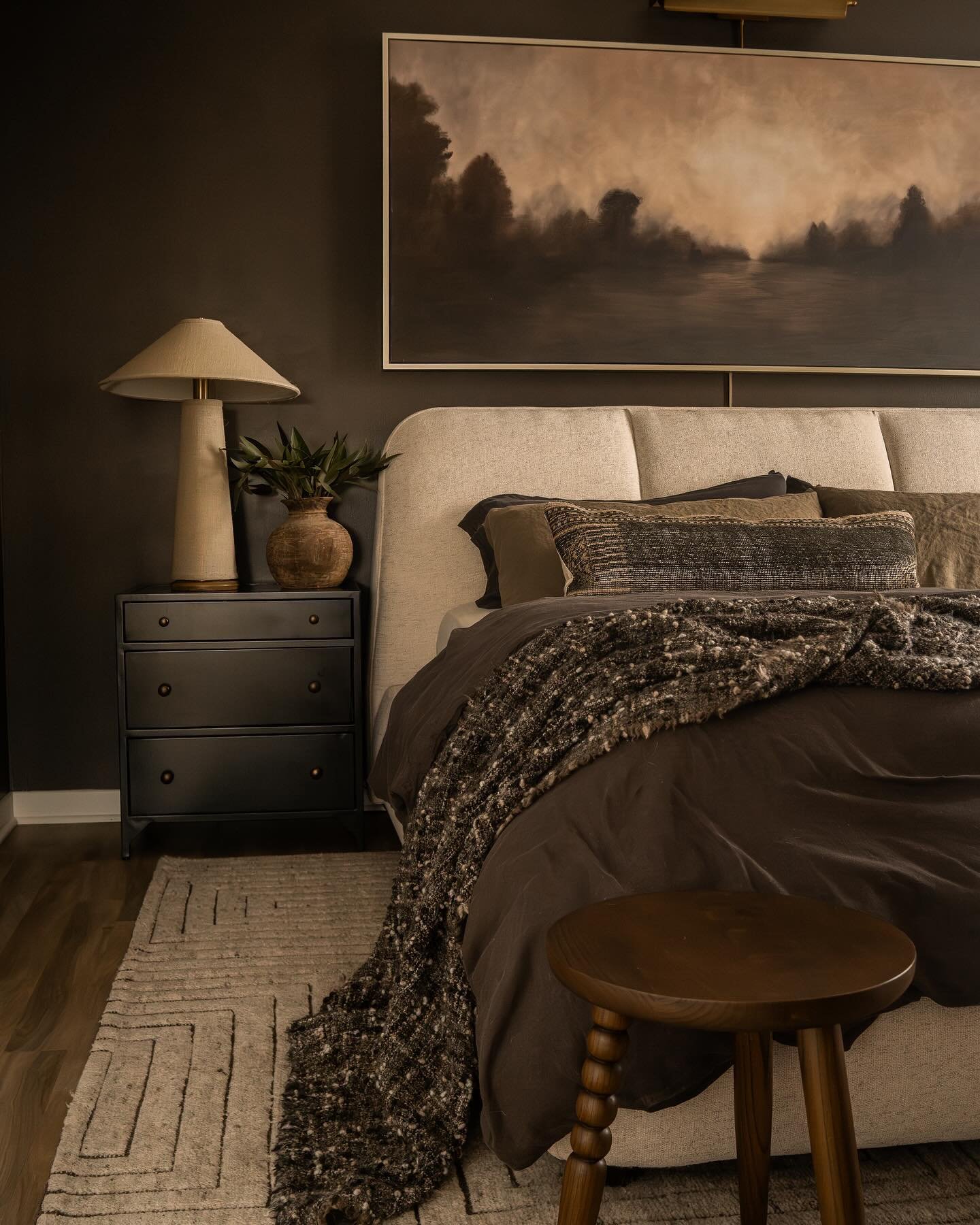 A small, neutral area rug with a subtle geometric pattern is placed next to a bed with dark bedding and a black nightstand.