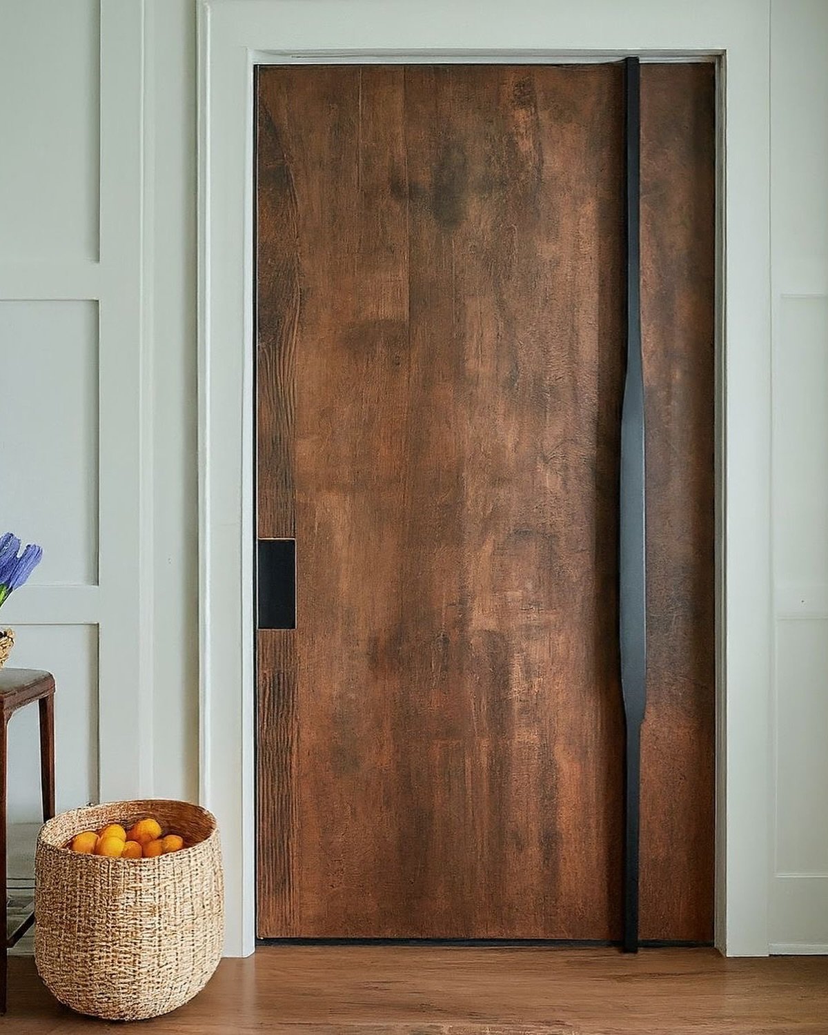 A close-up of a rustic, textured wooden door with an oxidized copper finish, featuring a unique, long, sculptural black handle and a clean white frame.