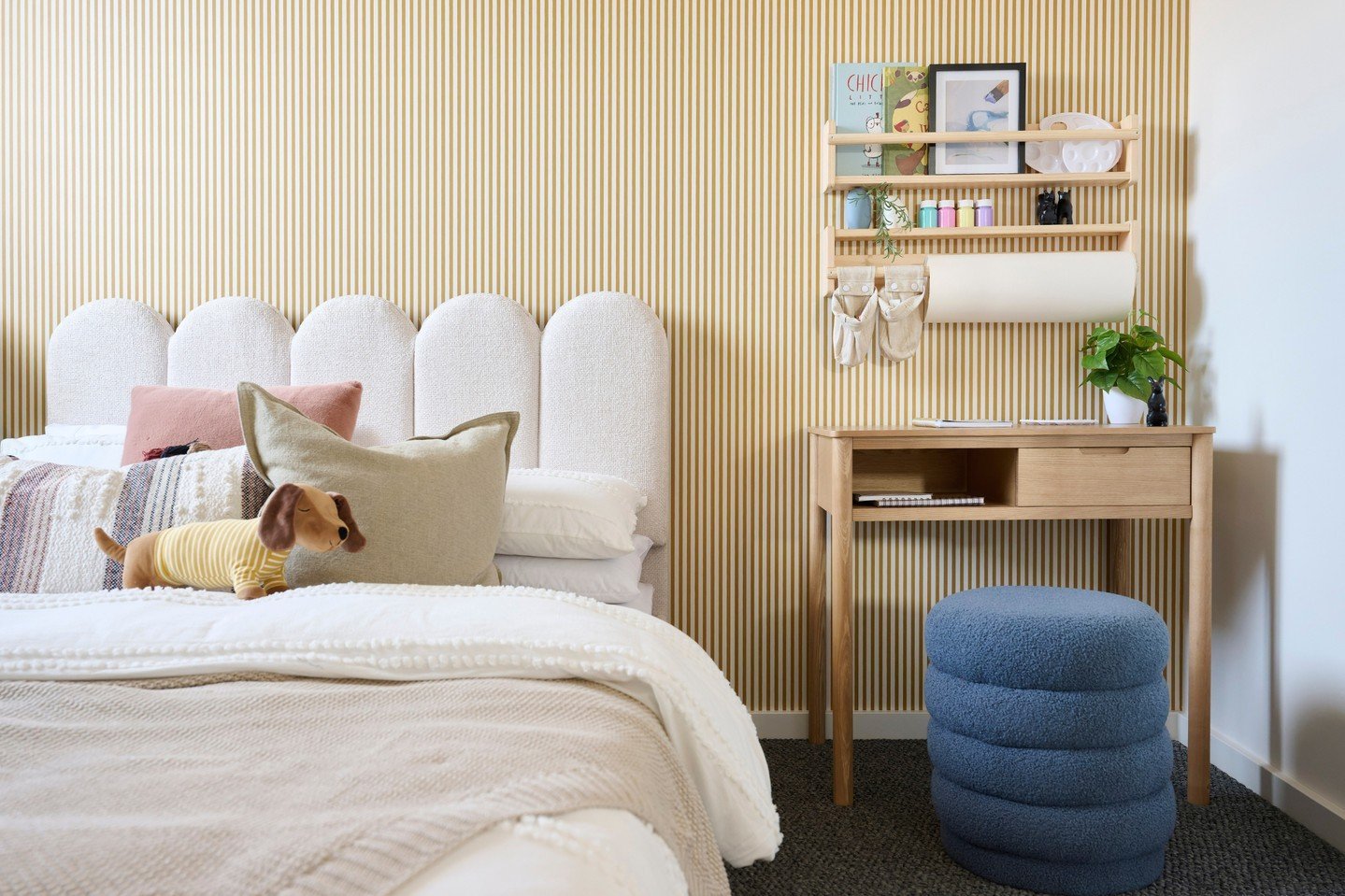 A cozy girl's bedroom with a white upholstered headboard with a scallop design, yellow striped wallpaper, and a wooden desk with a large paper roll holder.