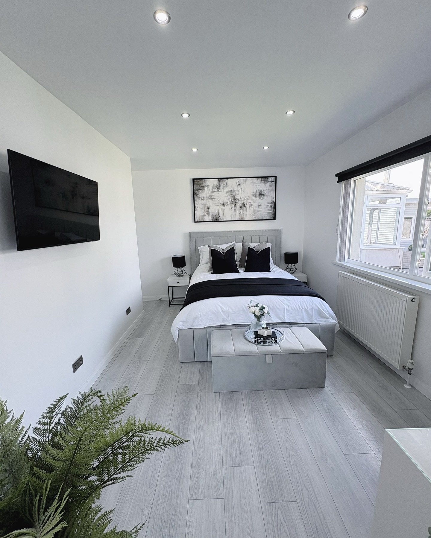 A modern bedroom with white walls, a light gray upholstered bed with black and white pillows, an abstract black and white painting, and a plush gray bench at the foot of the bed.