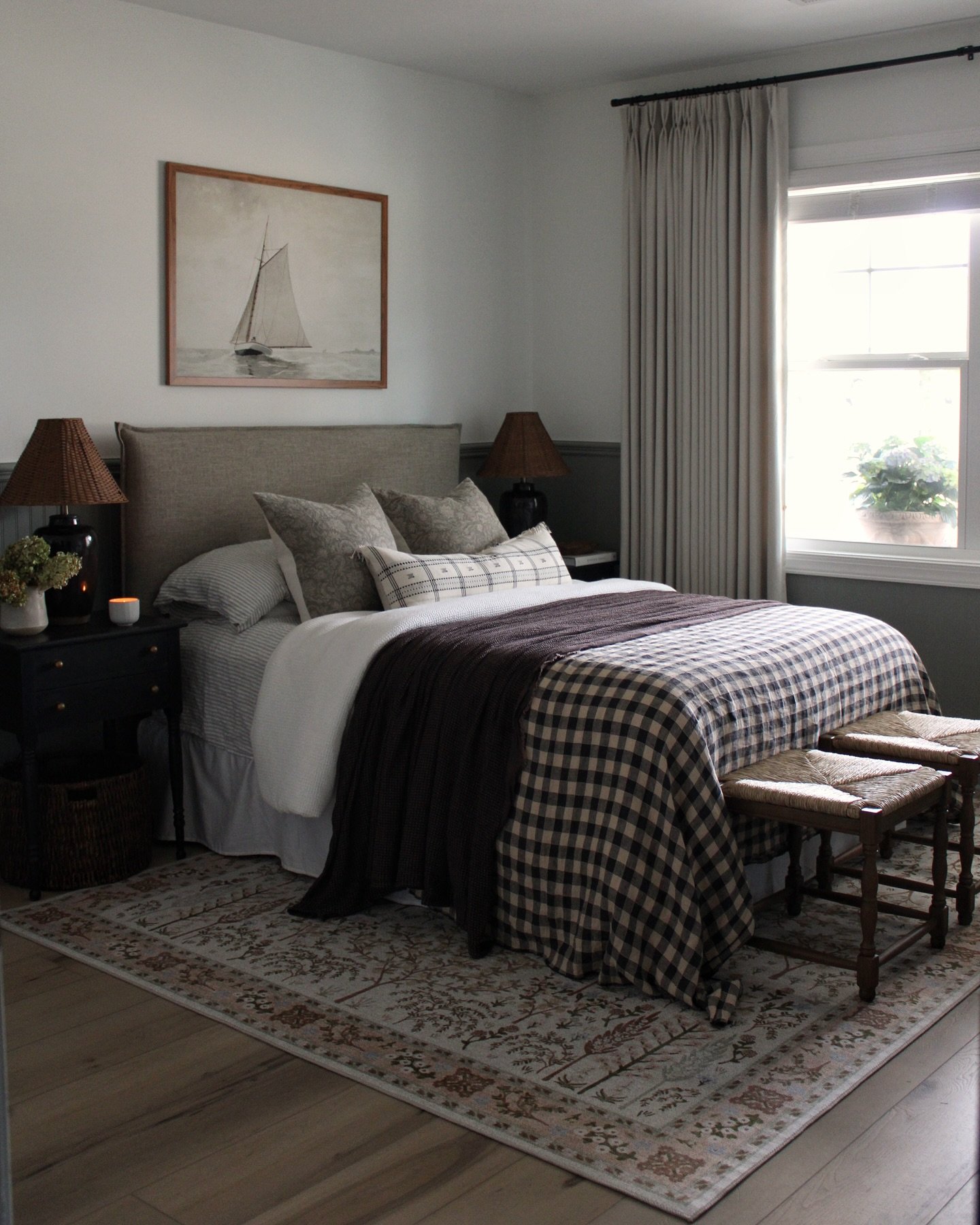 A cozy farmhouse guest bedroom with a queen-sized bed, gingham bedding, and a light-toned upholstered headboard. The room features a nautical painting, dark nightstands, and two small woven stools at the foot of the bed.