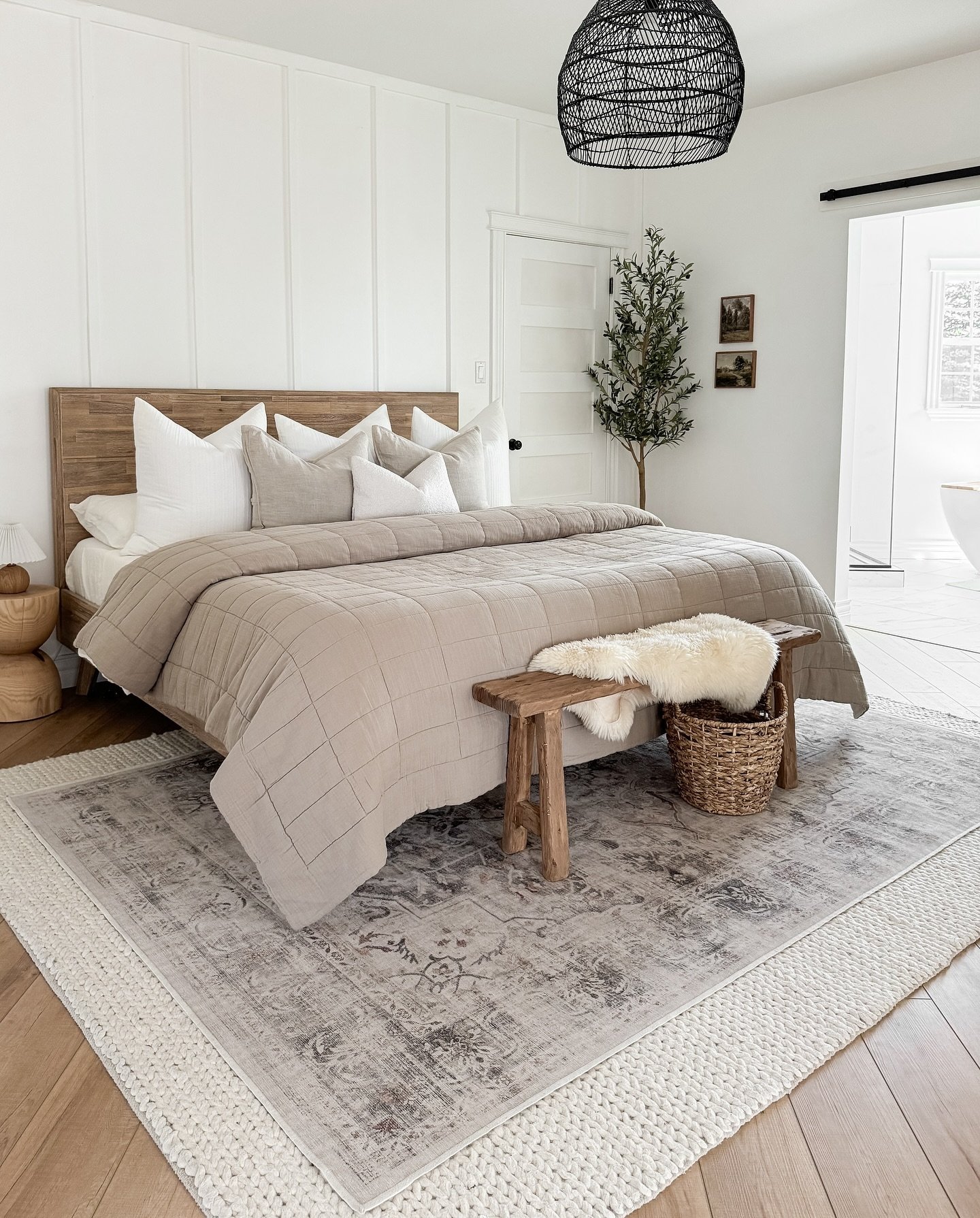 A bright bedroom with white paneled walls and a wooden headboard, with a bed dressed in beige bedding.