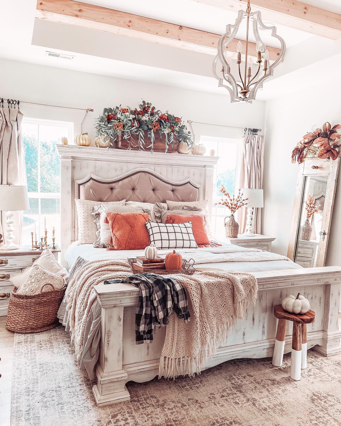 A bedroom with a distressed white bed, tufted headboard, and layered pillows in orange and plaid patterns.