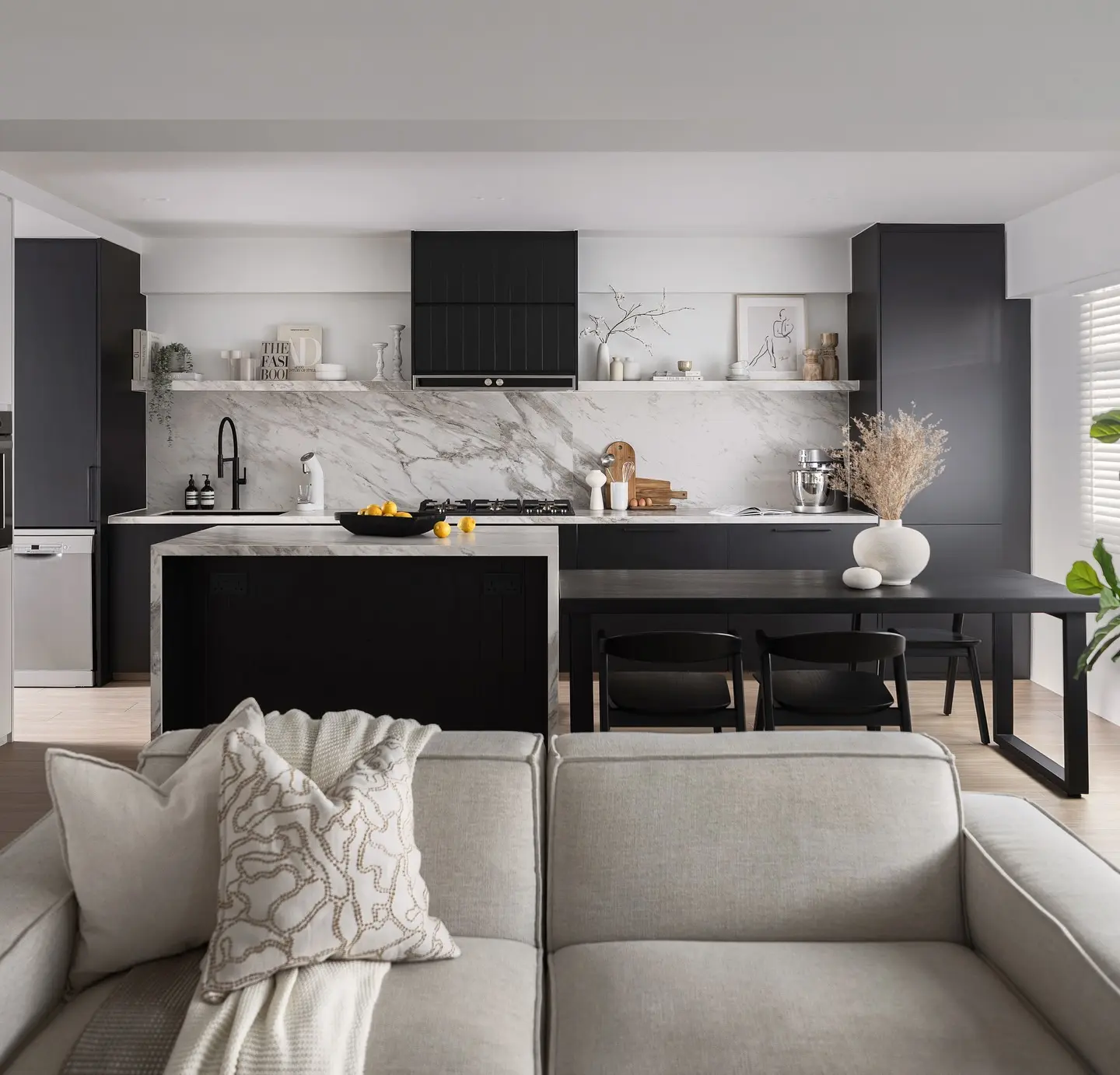A modern, open-concept kitchen and living room with a monochrome black and white color scheme. The kitchen has black and white cabinets, a black island, and a marble backsplash. A black dining table with chairs sits adjacent to the island, and a light-colored sofa is in the foreground.