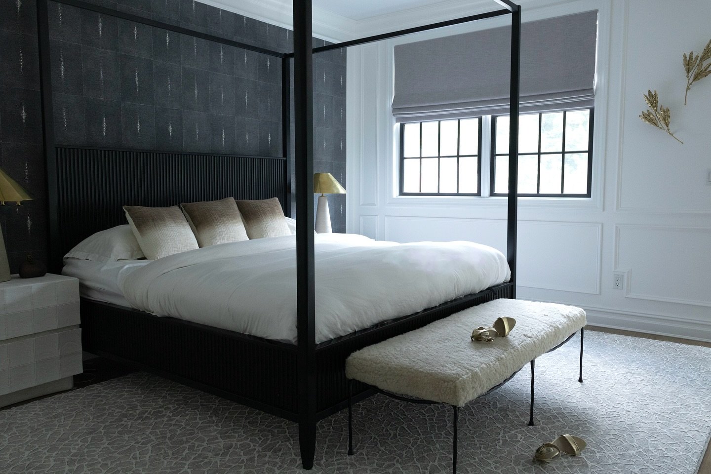 An elegant black and white bedroom with a black four-poster bed, patterned dark gray wallpaper, white wainscoting, and a white furry bench at the foot of the bed.