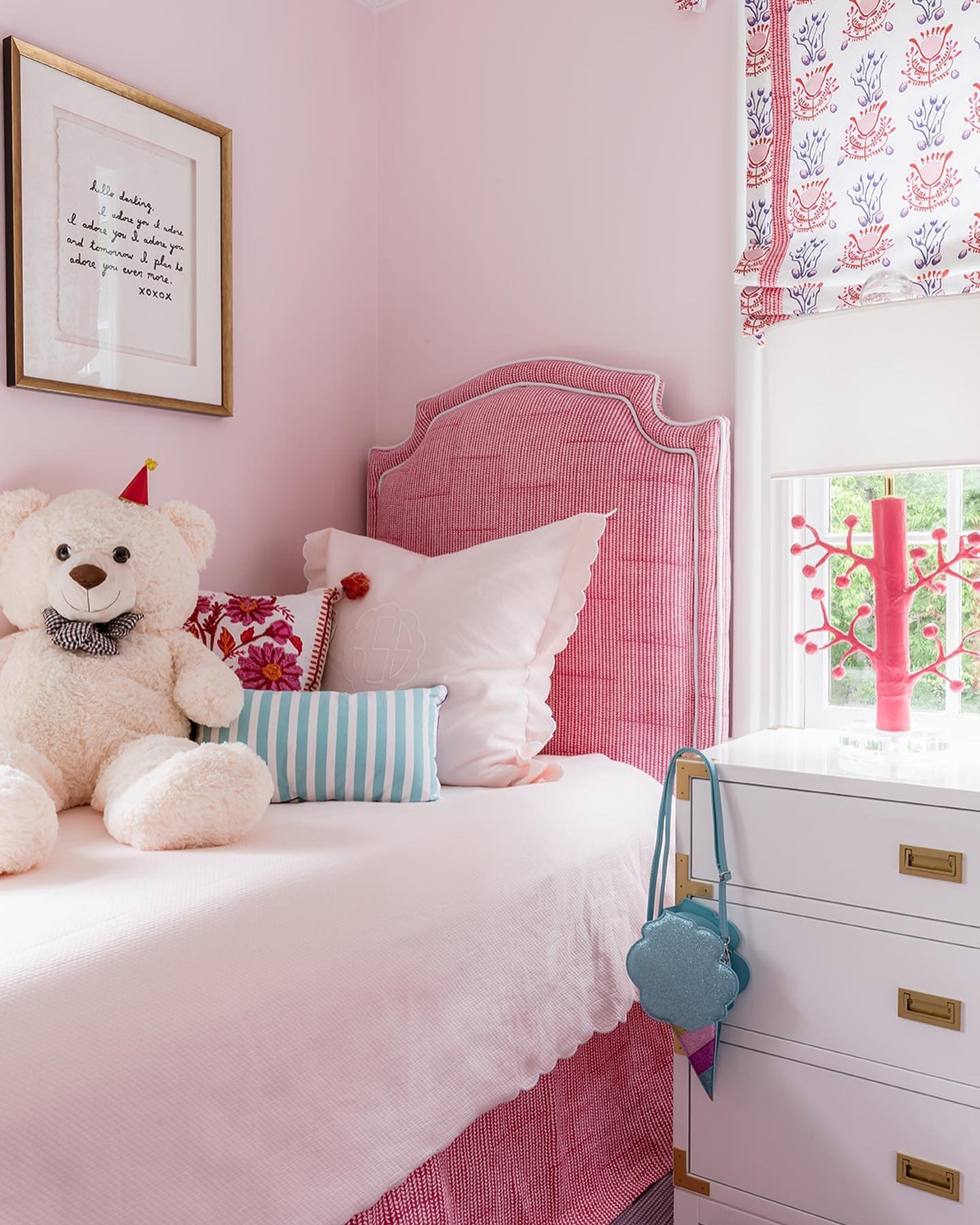 A sweet girl's bedroom with a pink-striped upholstered headboard, a large teddy bear, and a white nightstand with gold hardware.