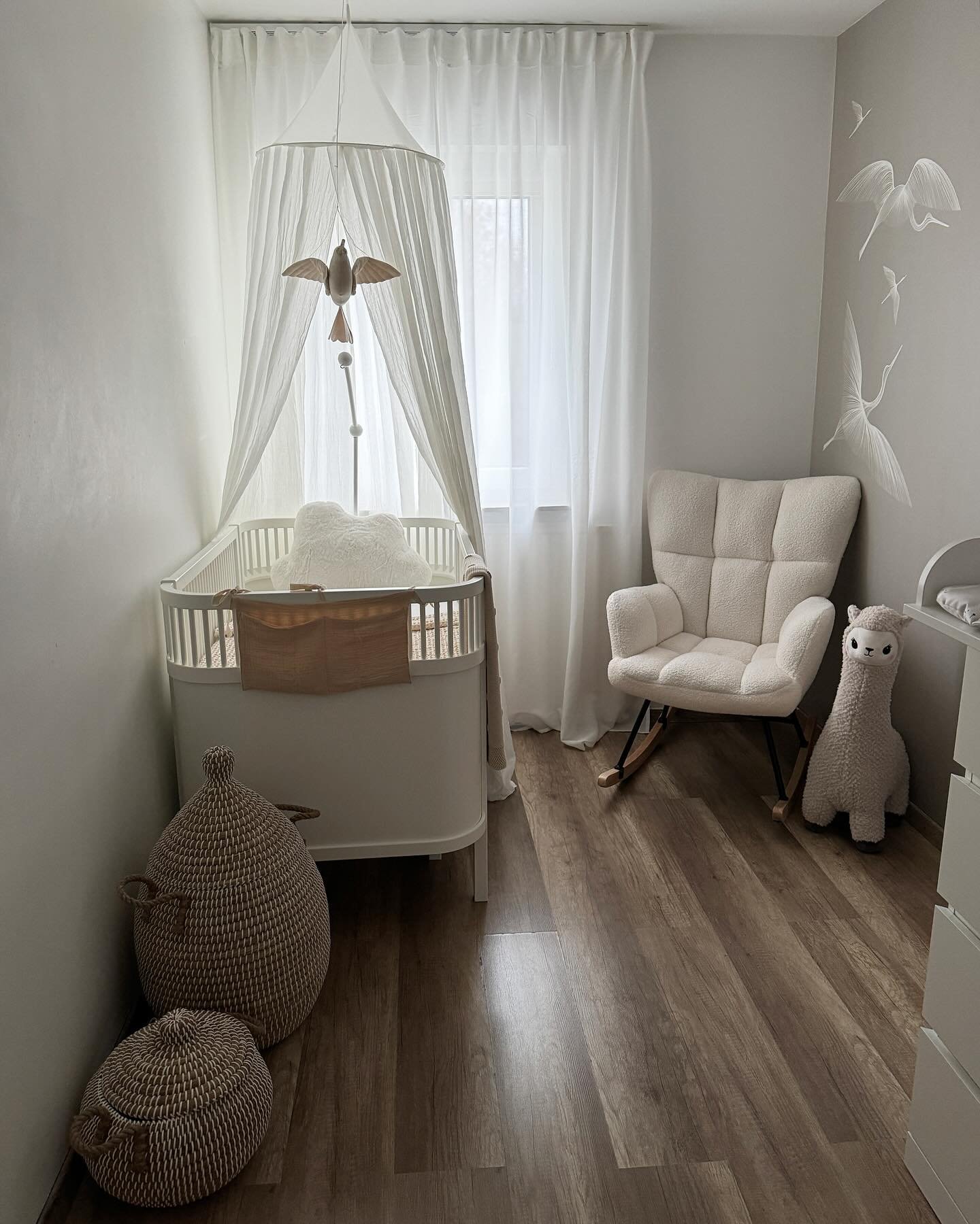 A bright minimalist nursery with a white crib and canopy, a teddy rocker chair, and bird wall decals.