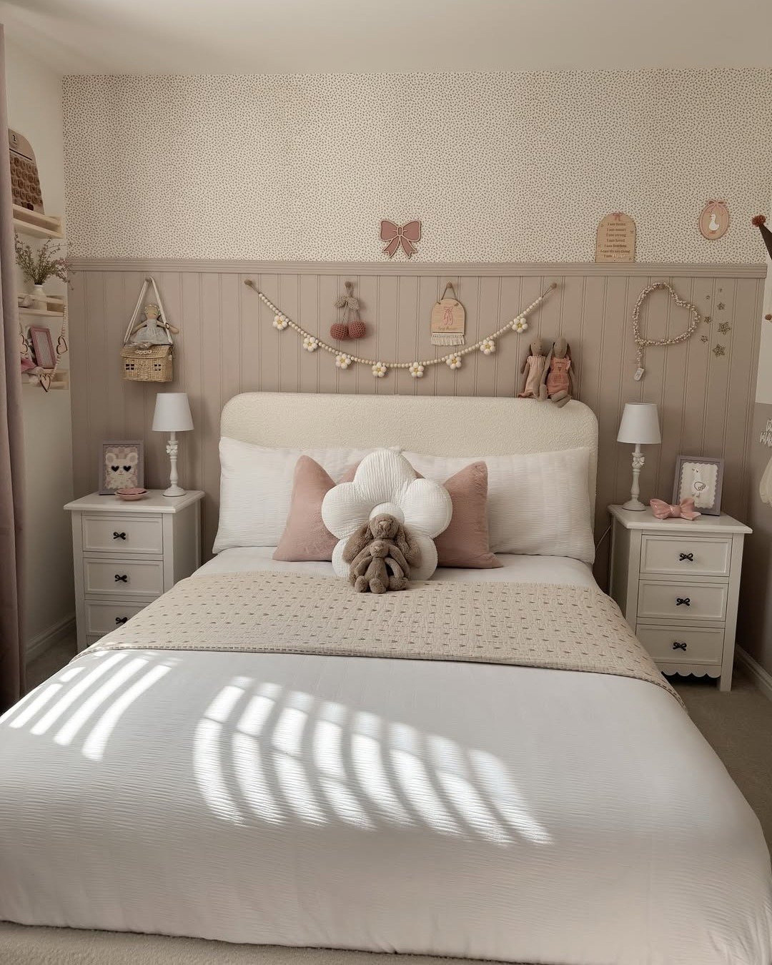 A cozy bedroom with half-paneled beige walls, small-print wallpaper, a floral garland, and white cottage-style nightstands.
