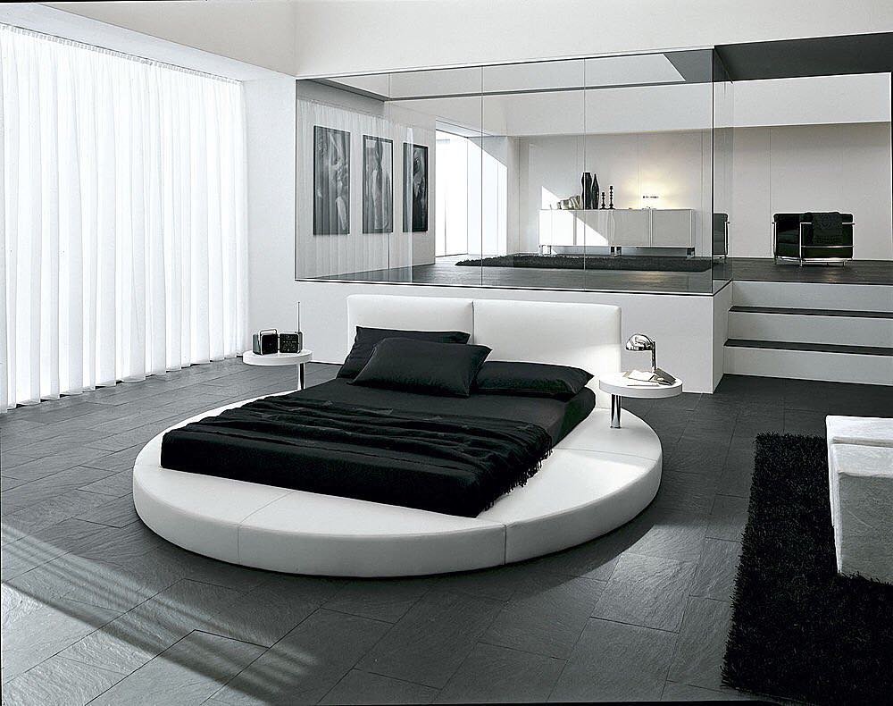  An ultra-modern bedroom with a circular white platform bed, black bedding, dark gray tile floors, a sunken level separated by a glass wall, and sheer white curtains.
