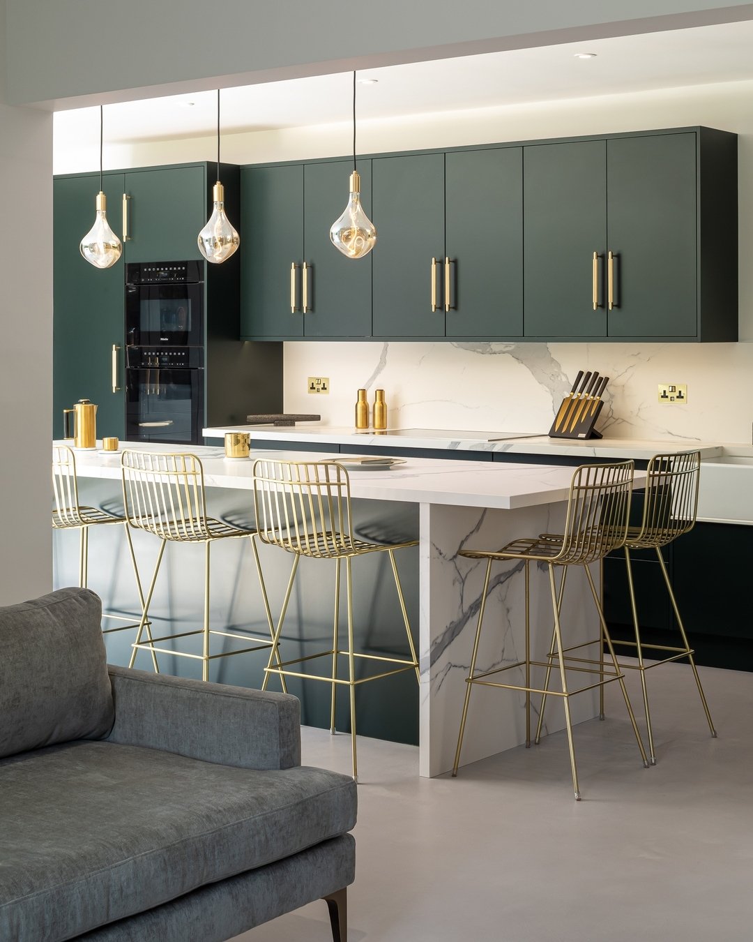 A small kitchen island with a marble waterfall countertop, four gold wire bar stools, and a dark green kitchen in a modern apartment.