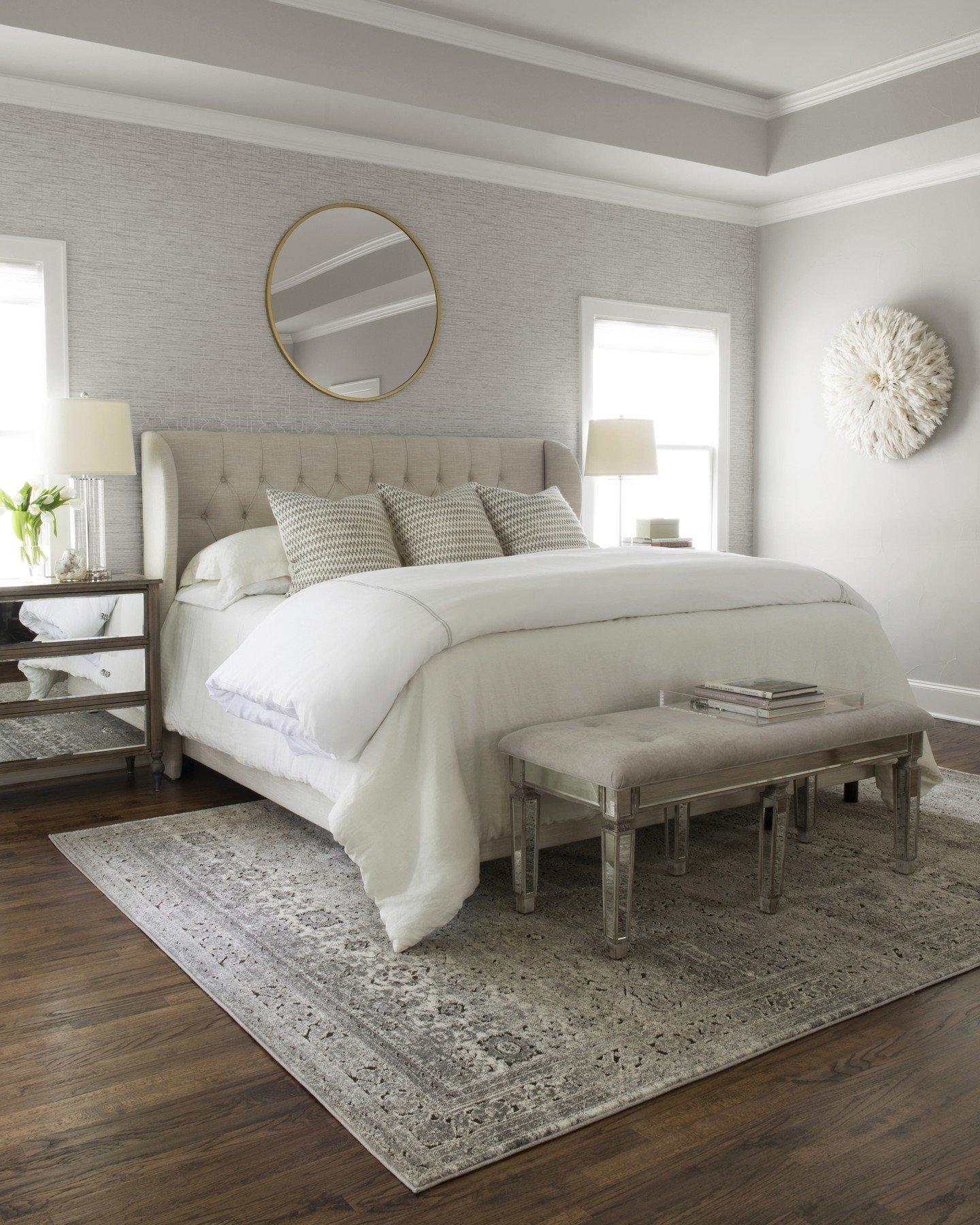 A traditional gray and white patterned area rug placed under a bed with a light-colored tufted headboard on a dark wood floor.