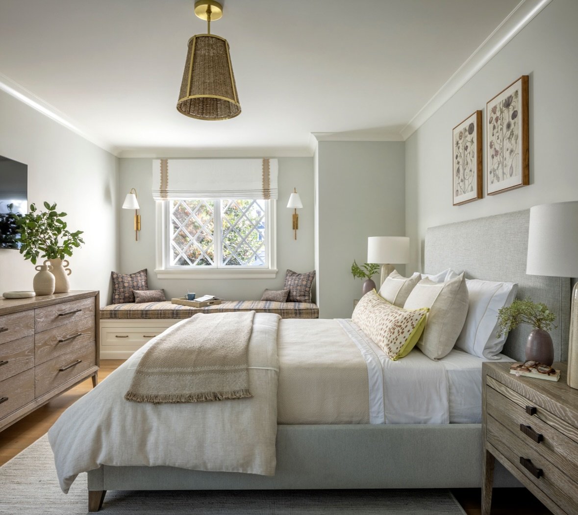 A tranquil and elegant guest bedroom with a gray upholstered bed, white and cream linens, and a built-in window seat. The room features textured wood furniture, botanical prints, and a rattan pendant light.