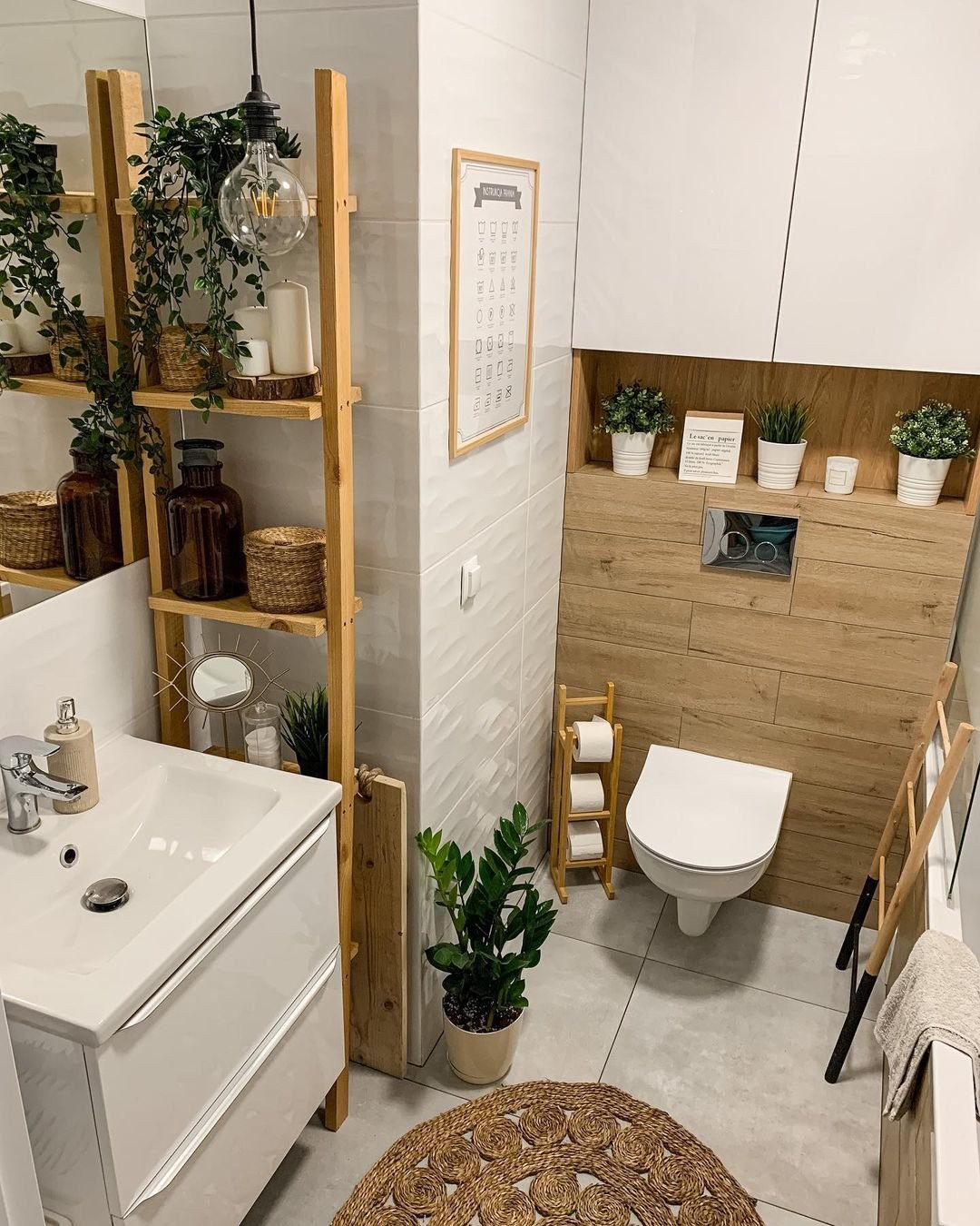 A small, bright bathroom with a rustic, bohemian style. It features a wall-mounted ladder shelf with plants, a wood-paneled toilet area, and a white floating vanity.