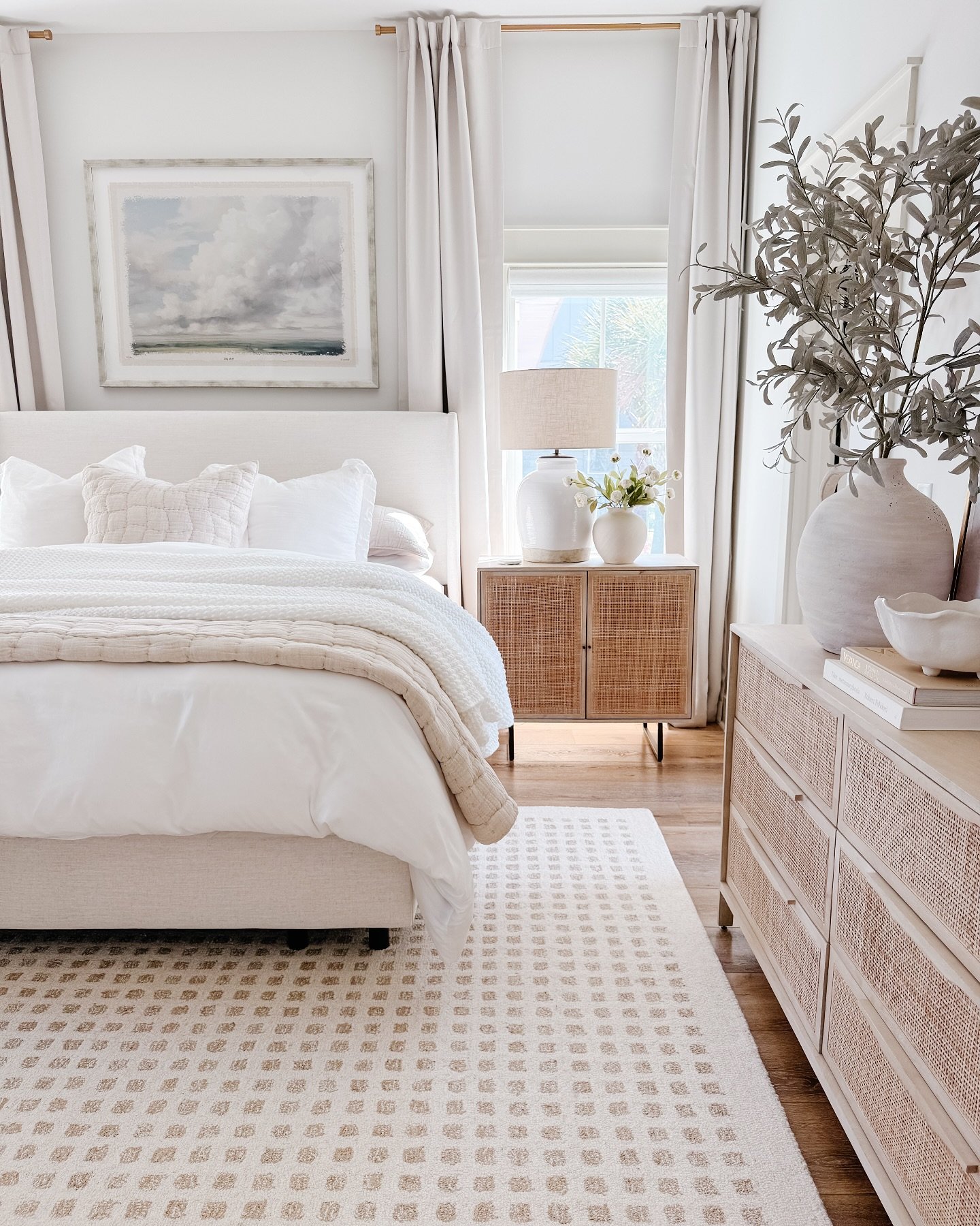 A neutral area rug with a small, repeating geometric pattern is placed under a light-colored bed with white bedding and a beige blanket.