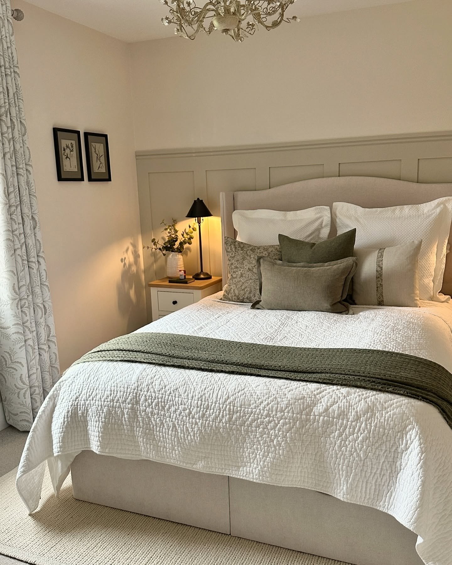 A cozy and elegant guest bedroom with a light gray upholstered bed, crisp white bedding, and soft green accent pillows and throws. The room features a wainscoting accent wall, a small white nightstand, and a vintage-style chandelier.