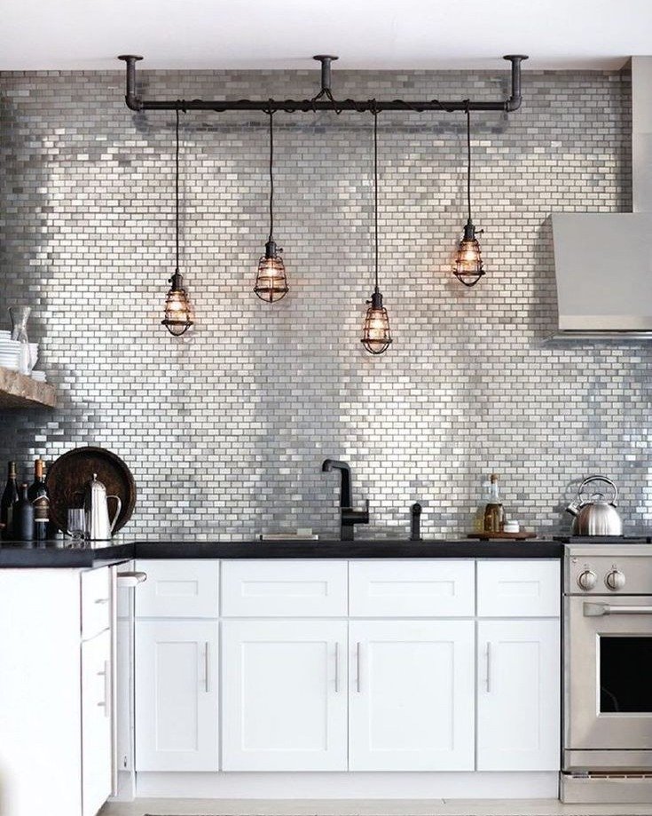 A kitchen featuring a backsplash of shimmering metallic mosaic tiles in a brick pattern, with a black countertop, white cabinets, and industrial cage pendant lights.