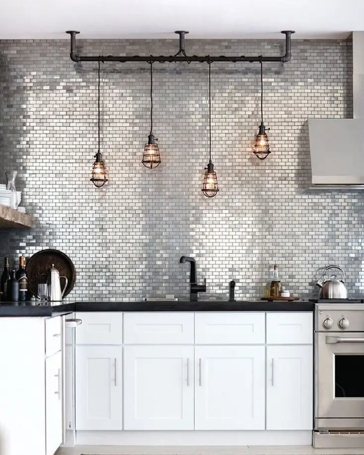 A kitchen featuring a backsplash of shimmering metallic mosaic tiles in a brick pattern, with a black countertop, white cabinets, and industrial cage pendant lights.