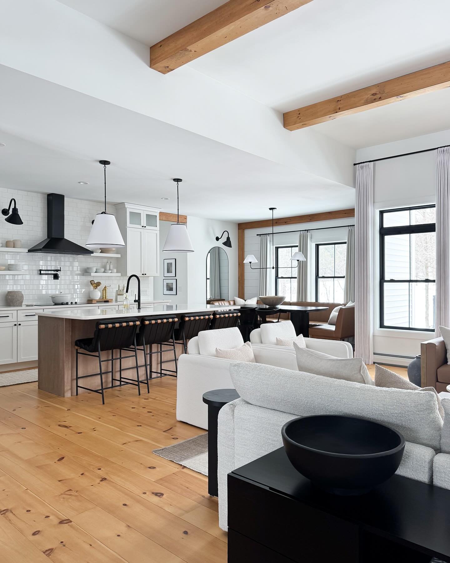 A bright and spacious open-concept kitchen and living room with a rustic farmhouse aesthetic. The kitchen has a white subway tile backsplash, a large island with black bar stools, and white cabinetry. The living area features a light grey sofa and wooden ceiling beams.
