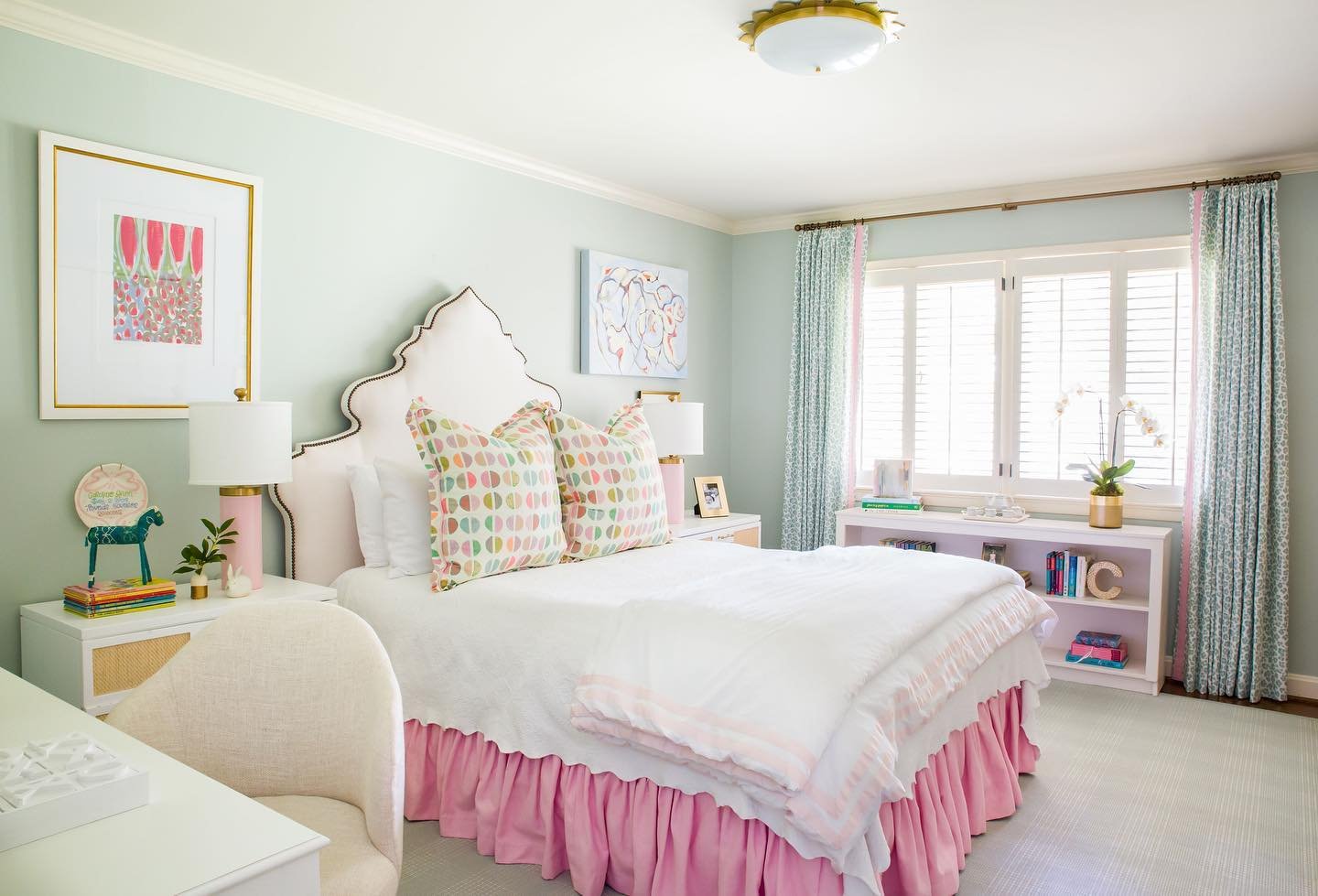 A bright bedroom with pale green walls, a white scalloped headboard, and vibrant pops of pink and multi-colored patterns in the decor.