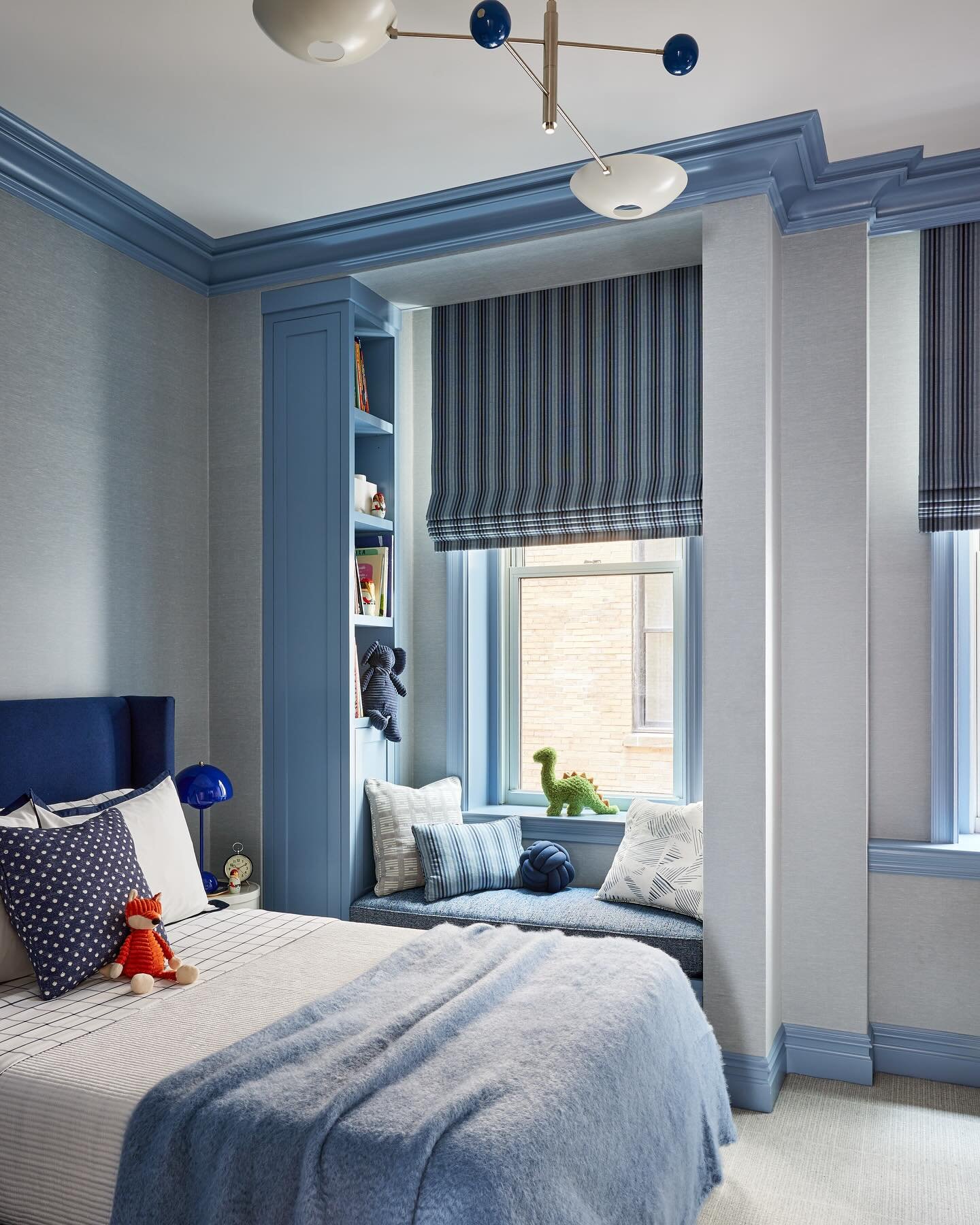 A teenage bedroom with a blue color scheme, featuring a window seat, built-in shelving, and a modern light fixture.