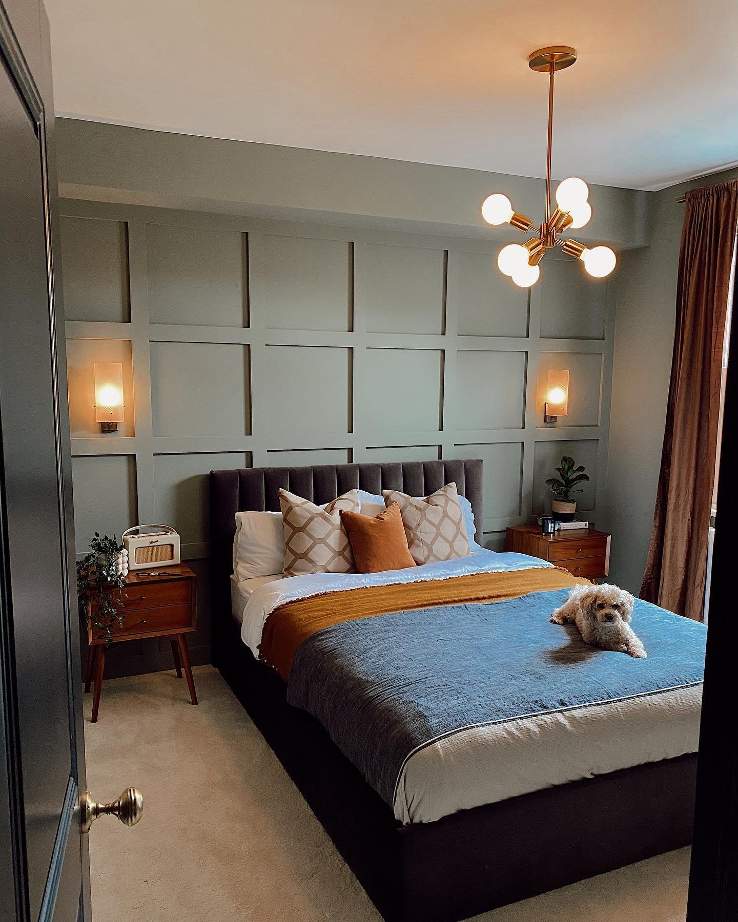 A modern guest bedroom with a gray upholstered bed and a paneled accent wall in a sage green color. The room features a dark blue and terracotta color scheme, mid-century modern nightstands, and a brass Sputnik-style chandelier.