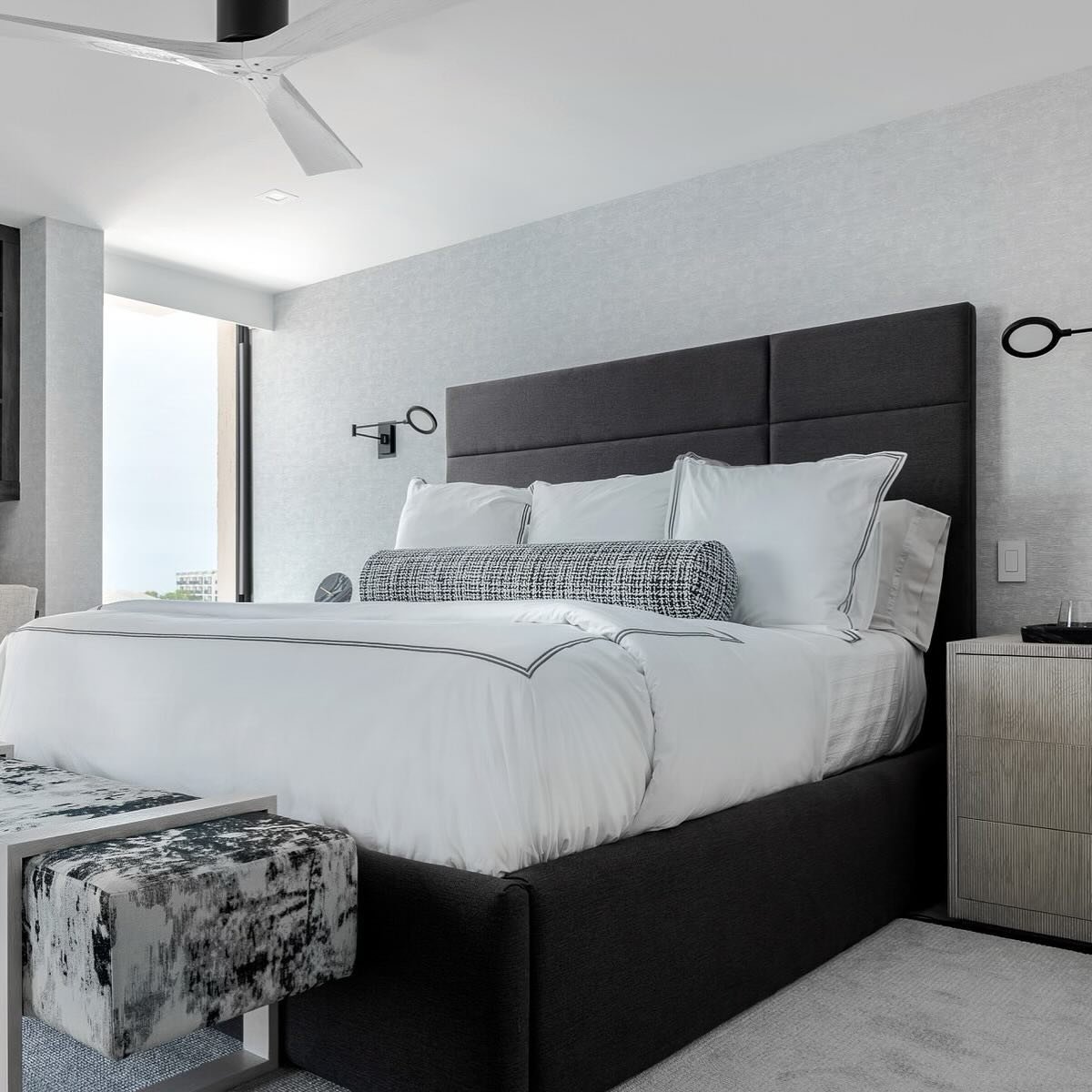 A contemporary bedroom with a dark gray paneled headboard, a bed with clean white bedding, a patterned bolster pillow, and a matching patterned bench at the foot of the bed.