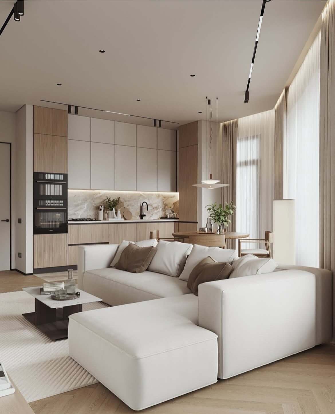 A modern, minimalist open-concept living and kitchen area. A large white sectional sofa sits on a textured rug in the foreground. The kitchen features flat-panel light wood and grey cabinetry with a marble backsplash. A small round dining table with two chairs is visible next to a floor lamp.