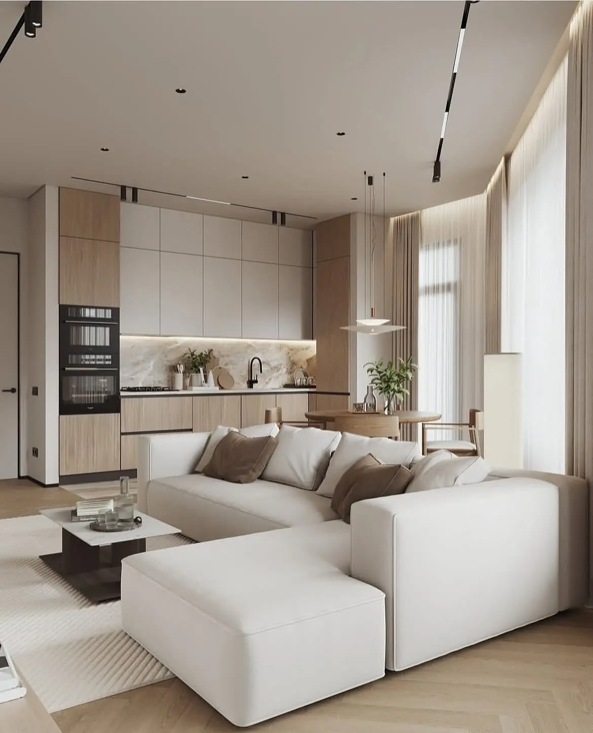 A modern, minimalist open-concept living and kitchen area. A large white sectional sofa sits on a textured rug in the foreground. The kitchen features flat-panel light wood and grey cabinetry with a marble backsplash. A small round dining table with two chairs is visible next to a floor lamp.